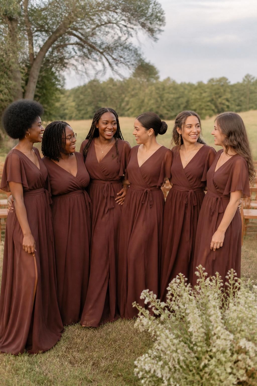 Realistic countryside wedding photograph of six bridesmaids wearing mahogany brown chiffon wrap dresses with crossed V-necklines, adjustable waist ties, flutter sleeves, and flowing floor-length skirts, women with deep ebony, rich brown, medium brown, golden tan, olive, and fair skin tones, varied ages, bust sizes, heights, and body shapes represented naturally, bright friendly faces, realistic skin texture and light makeup, hairstyles including a rounded afro, shoulder-length locs, long braids, smooth low bun, layered curls, and loose waves, standing in an open meadow ceremony space with cream wildflowers, wooden chairs, mature trees, and a soft late-afternoon sky, light wind moving the sleeves and skirts, authentic chiffon transparency and candid group interaction, human DSLR photo-realistic style.