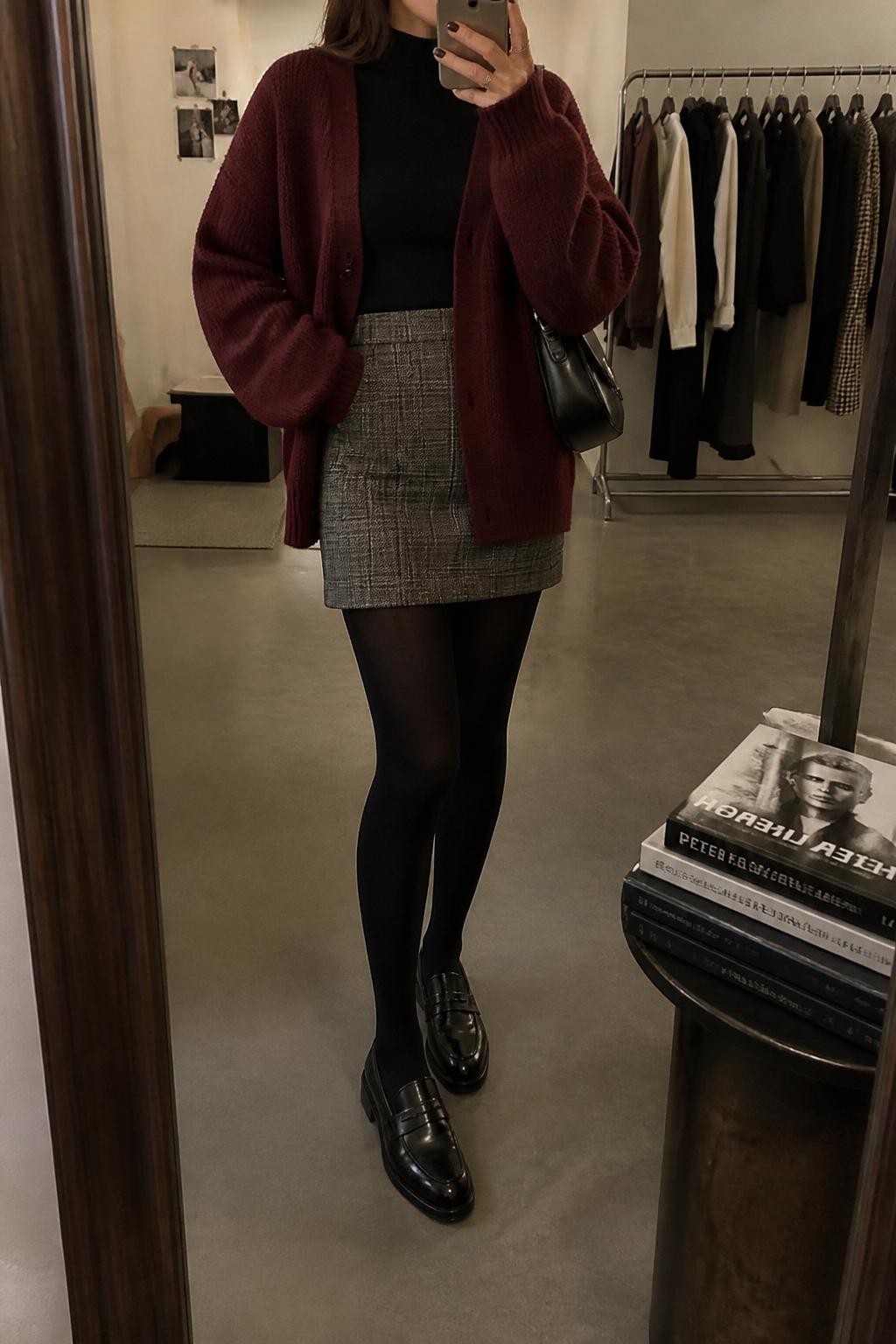 Mirror selfie of a woman wearing a burgundy oversized knit cardigan, black fitted turtleneck, gray plaid wool mini skirt, black opaque tights, black leather loafers, and a small black shoulder bag, holding a phone in front of a mirror, cropped at NECK so the face does not show, relaxed casual selfie pose, in a fashion studio mirror corner, warm moody lighting, with a clothing rack with garments behind and a stack of fashion magazines on a side table, latest iPhone photo quality.