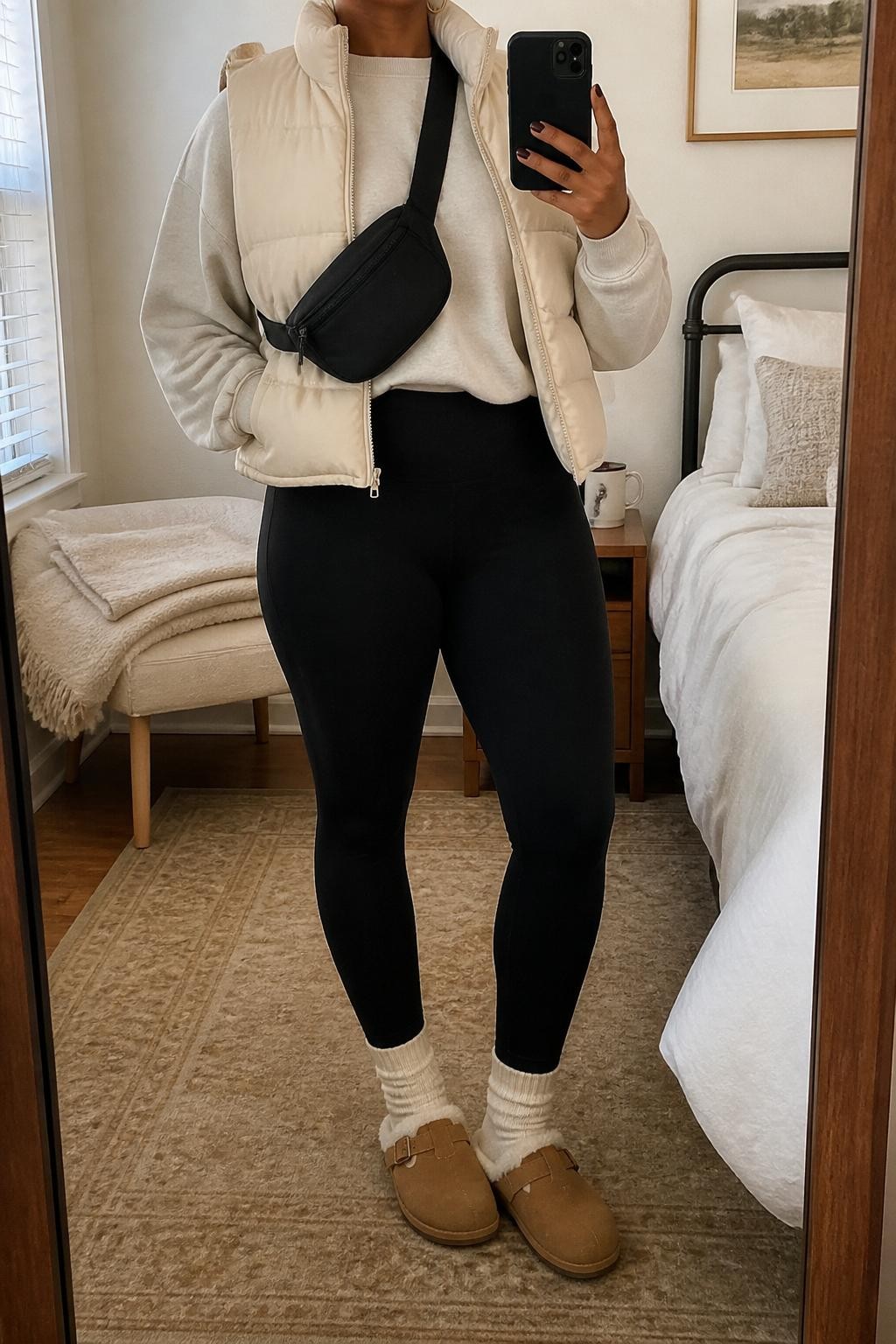 mirror selfie of a Black woman wearing the exact outfit described above, clearly showing shearling-lined tan Birkenstock-style clogs with cream socks, black high-waist leggings, an oversized oatmeal sweatshirt, a cream puffer vest, a black belt bag worn crossbody, and small gold hoop earrings, holding a phone in front of a mirror, cropped at NECK so the face does not show, with a relaxed casual selfie pose, in a cozy bedroom mirror setup, natural window light, with a folded blanket on a chair and a coffee mug on a bedside table, latest iPhone photo quality.