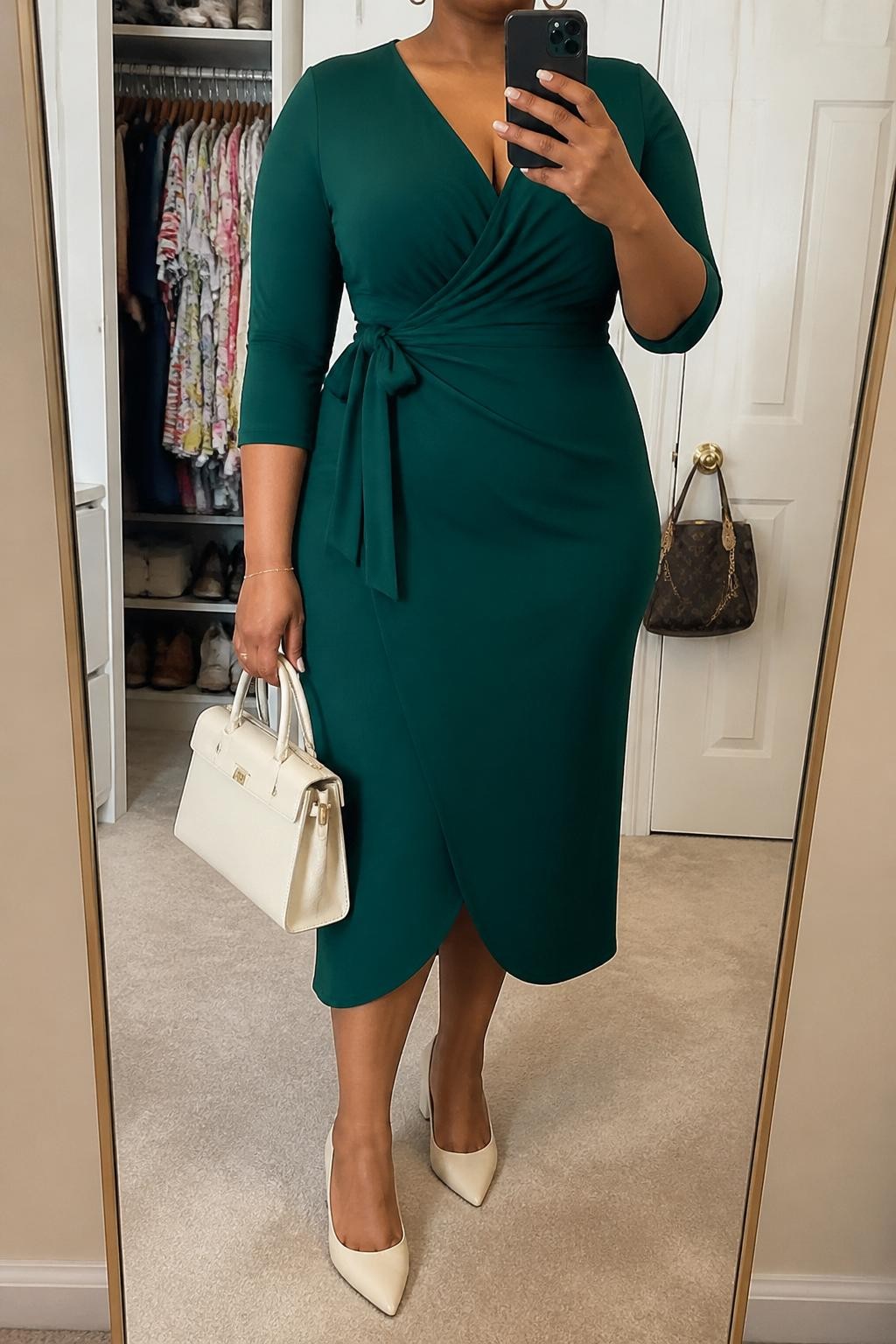 Mirror selfie of a plus size woman wearing an emerald green crepe wrap midi dress with three-quarter sleeves, a modest V-neckline, and a matching waist tie, cream pointed block heels, a structured cream handbag, and delicate gold hoop earrings, clearly showing the exact dress, emerald crepe, wrap shape, cream footwear, and accessories listed, holding a phone in front of a mirror, cropped at the neck so her face does not appear, relaxed graceful selfie stance, in a walk-in closet mirror with soft indoor lighting, an open closet with clothes visible and a handbag hanging on a door handle, realistic Sunday church outfit, latest iPhone photo quality.