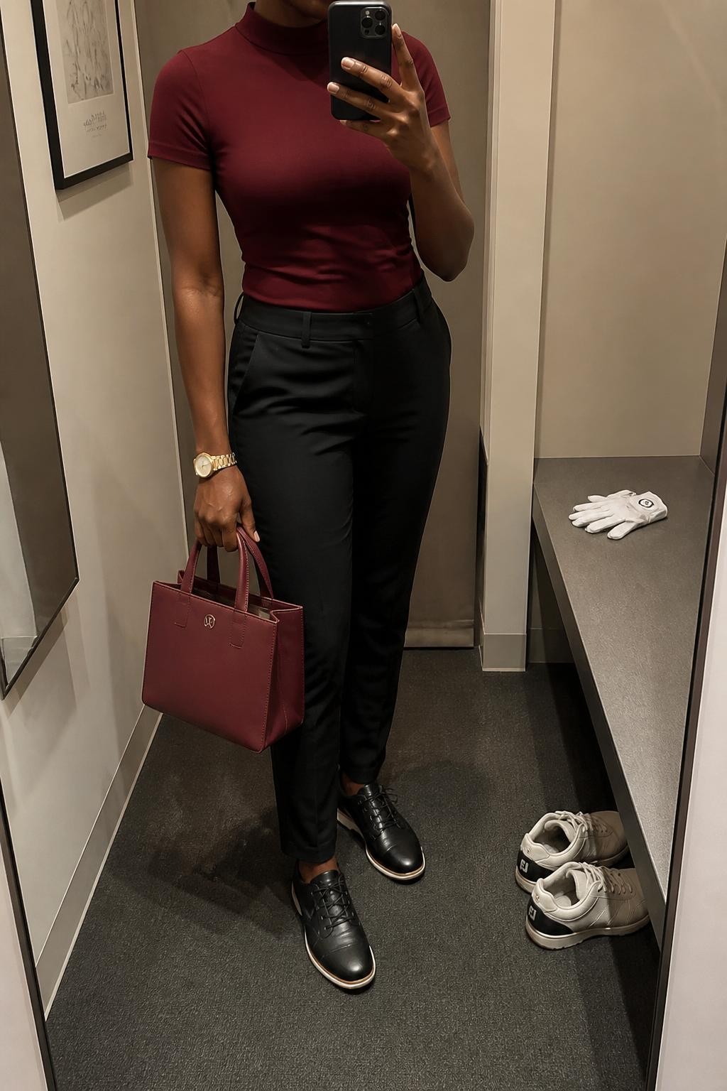 Mirror selfie of a Black woman wearing a burgundy fitted mock-neck golf top in stretch fabric tucked into black tapered golf trousers, clearly showing black leather spikeless golf shoes, a black visor, a structured burgundy mini tote, and a slim gold wristwatch, holding an iPhone in front of a minimal dressing room mirror, cropped at the neck so the face does not show, relaxed casual selfie pose, warm moody lighting, a golf glove on the counter and a pair of shoes on the floor nearby, natural unstaged setting, latest iPhone photo quality.