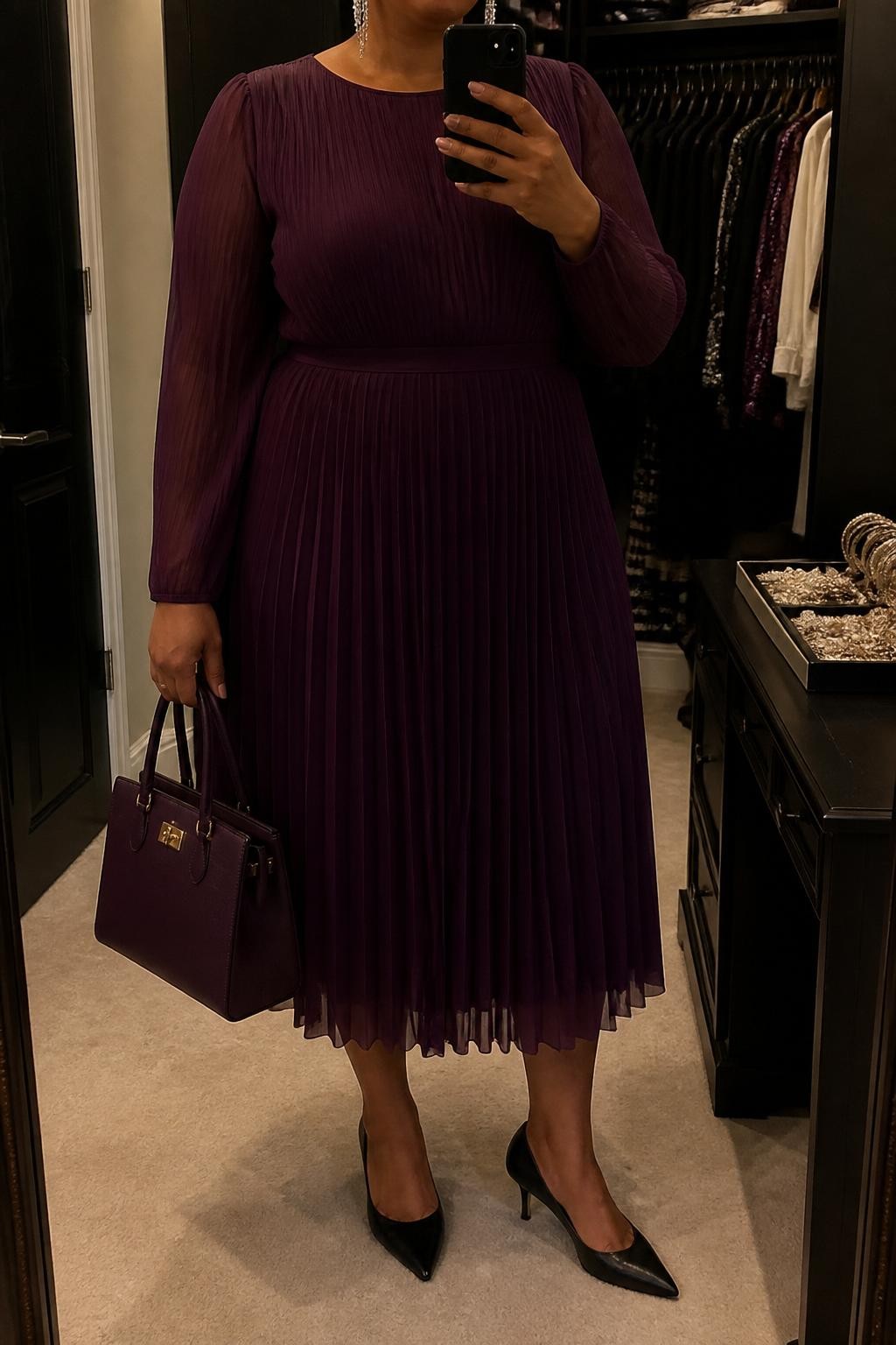 Mirror selfie of a plus size woman wearing a plum purple pleated midi dress with long sheer sleeves over an opaque lining, a modest round neckline, and a softly fitted waist, black pointed kitten heels, a structured plum handbag, and silver crystal drop earrings, clearly showing the exact dress, plum pleats, black footwear, handbag, and accessories listed, holding a phone in front of a mirror, cropped at the neck so the face does not show, relaxed graceful selfie pose, in a walk-in closet mirror with warm moody lighting, an open closet with clothes visible and a jewelry tray on the vanity, realistic special church service outfit, latest iPhone photo quality.
