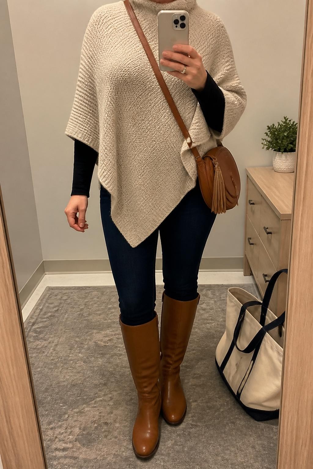 Mirror selfie of a curvy woman wearing an oatmeal knit poncho, black fitted long-sleeve top, dark indigo skinny jeans, tan riding boots, and a brown crossbody bag, holding a phone in front of a mirror, cropped at NECK so the face does not show, relaxed casual selfie pose, in a minimal dressing room, warm indoor lighting, with a tote bag resting on the floor and a small plant on a dresser, latest iPhone photo quality.
