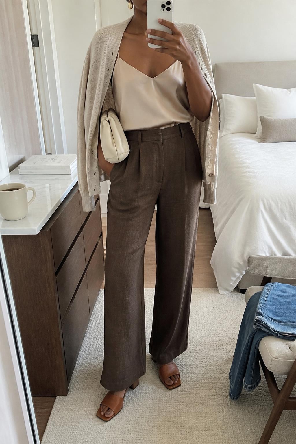 mirror selfie of a Black woman wearing the exact outfit described above, clearly showing espresso brown linen trousers, champagne satin camisole, lightweight oatmeal cardigan draped over the shoulders, brown flat mules, a cream clutch, and gold drop earrings, holding a phone in front of a mirror, cropped at NECK so the face does not show, with a relaxed casual selfie pose, in a bright bedroom mirror setup, soft natural light, with folded denim on a chair and a coffee cup on a dresser, latest iPhone photo quality.