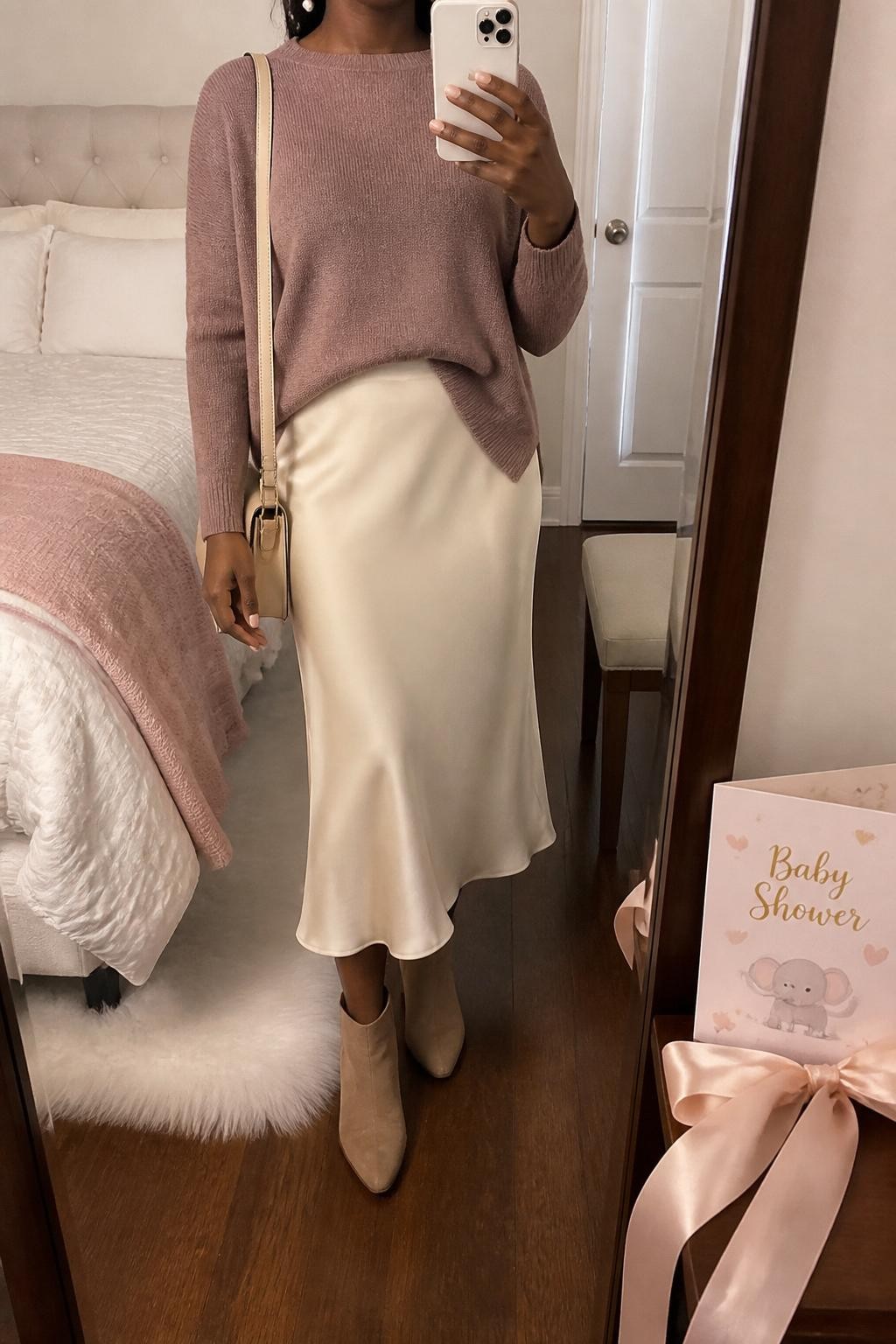 mirror selfie of a Black woman wearing the exact outfit described above, clearly showing a mauve soft knit sweater tucked slightly into a cream satin midi skirt, taupe suede ankle boots, a beige shoulder bag, and pearl drop earrings, holding a phone in front of a mirror, cropped at NECK so the face does not show, with a relaxed casual selfie pose, in a cozy bedroom mirror setup, warm indoor lighting, with a baby shower card and pastel ribbon on the vanity, latest iPhone photo quality.