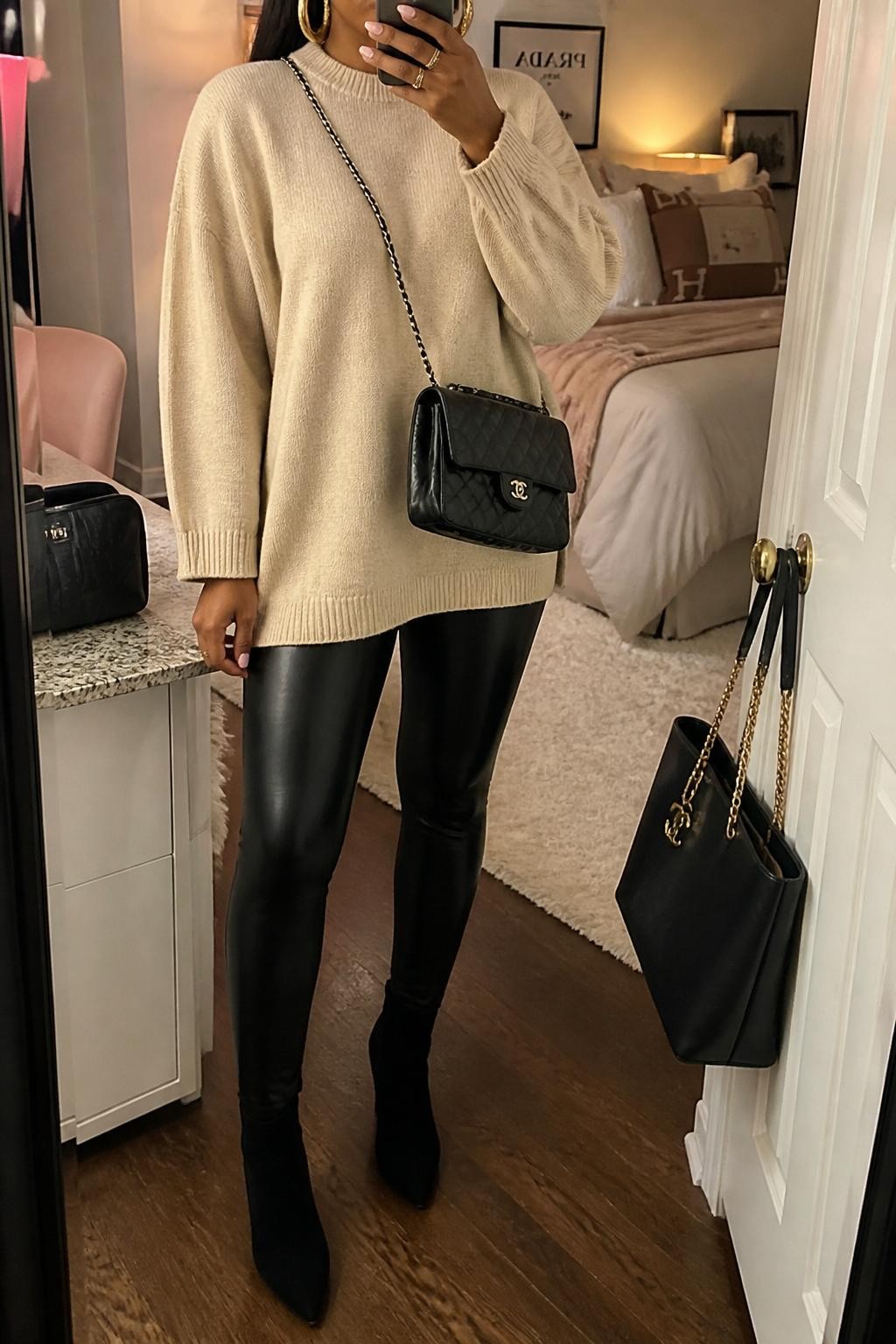 Mirror selfie of a Black woman wearing an oatmeal oversized sweater, black faux leather leggings, black suede ankle boots, gold hoop earrings, and a black quilted crossbody bag, holding a phone in front of a mirror, cropped at NECK so the face does not show, relaxed casual selfie pose, in a feminine-themed bedroom, evening golden hour, with a handbag hanging on a door handle and a cosmetic bag on the counter, latest iPhone photo quality.