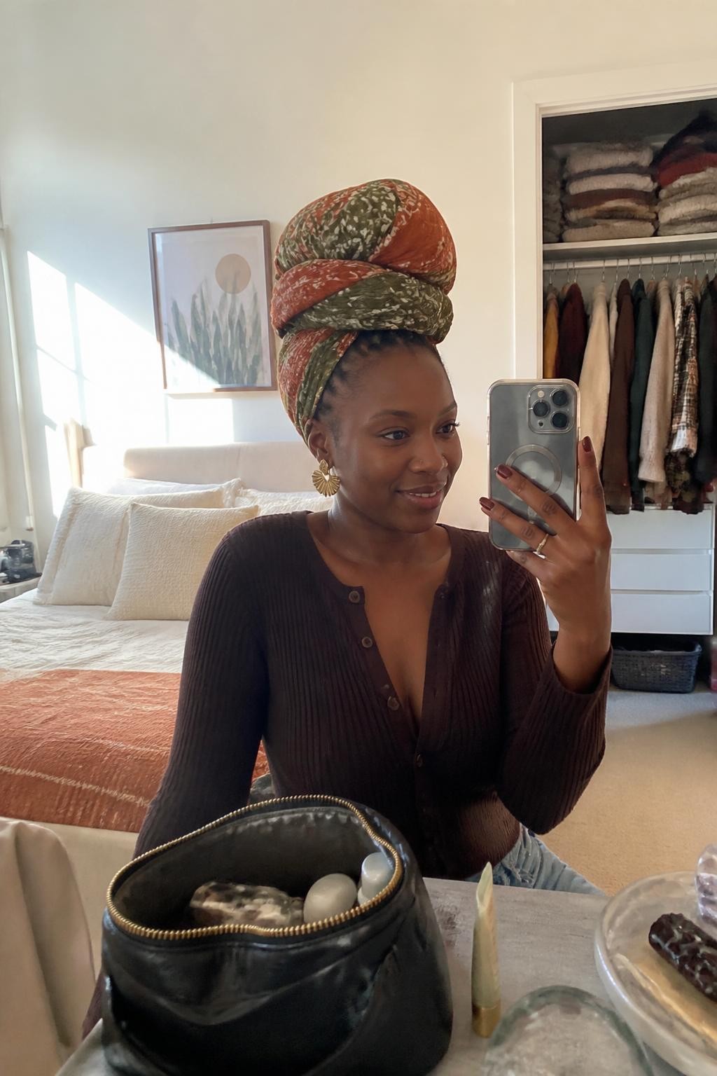 Image prompt: Photo of a Black woman wearing a tall rust-and-olive printed headwrap bun over hidden protective braids, candid mirror selfie, sun-drenched bedroom with folded sweaters and a slightly open closet behind her, natural window light, containing a makeup bag on the dresser and statement earrings peeking beneath the wrap, realistic relaxed fall morning moment, latest iPhone photo quality.