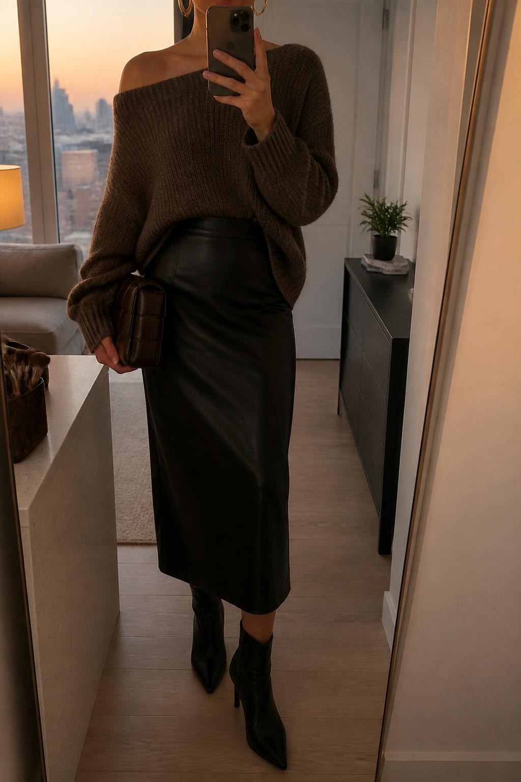 mirror selfie of a woman wearing the exact outfit described above, clearly showing a mocha off-shoulder sweater, black leather midi skirt, black heeled ankle boots, a dark brown clutch, and gold hoop earrings, holding a phone in front of a mirror, cropped at NECK so the face does not show, with a relaxed casual selfie pose, in a stylish city apartment, evening golden hour, with a makeup brush or cosmetic bag on the counter and a small plant on a dresser, latest iPhone photo quality.