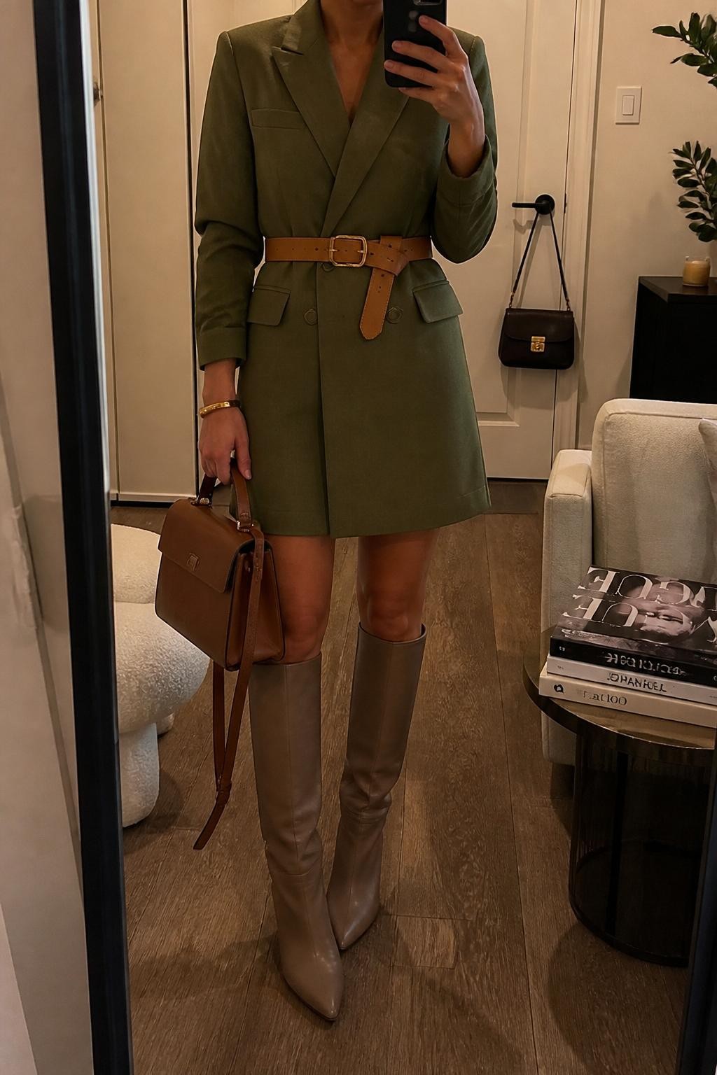 mirror selfie of a woman wearing the exact outfit described above, clearly showing an olive green blazer dress with a modest length, taupe knee-high boots, a tan leather belt, a brown structured satchel, and a gold bracelet, holding a phone in front of a mirror, cropped at NECK so the face does not show, with a relaxed casual selfie pose, in a stylish city apartment, warm moody lighting, with a handbag hanging on a door handle and a stack of fashion magazines on a side table, latest iPhone photo quality.