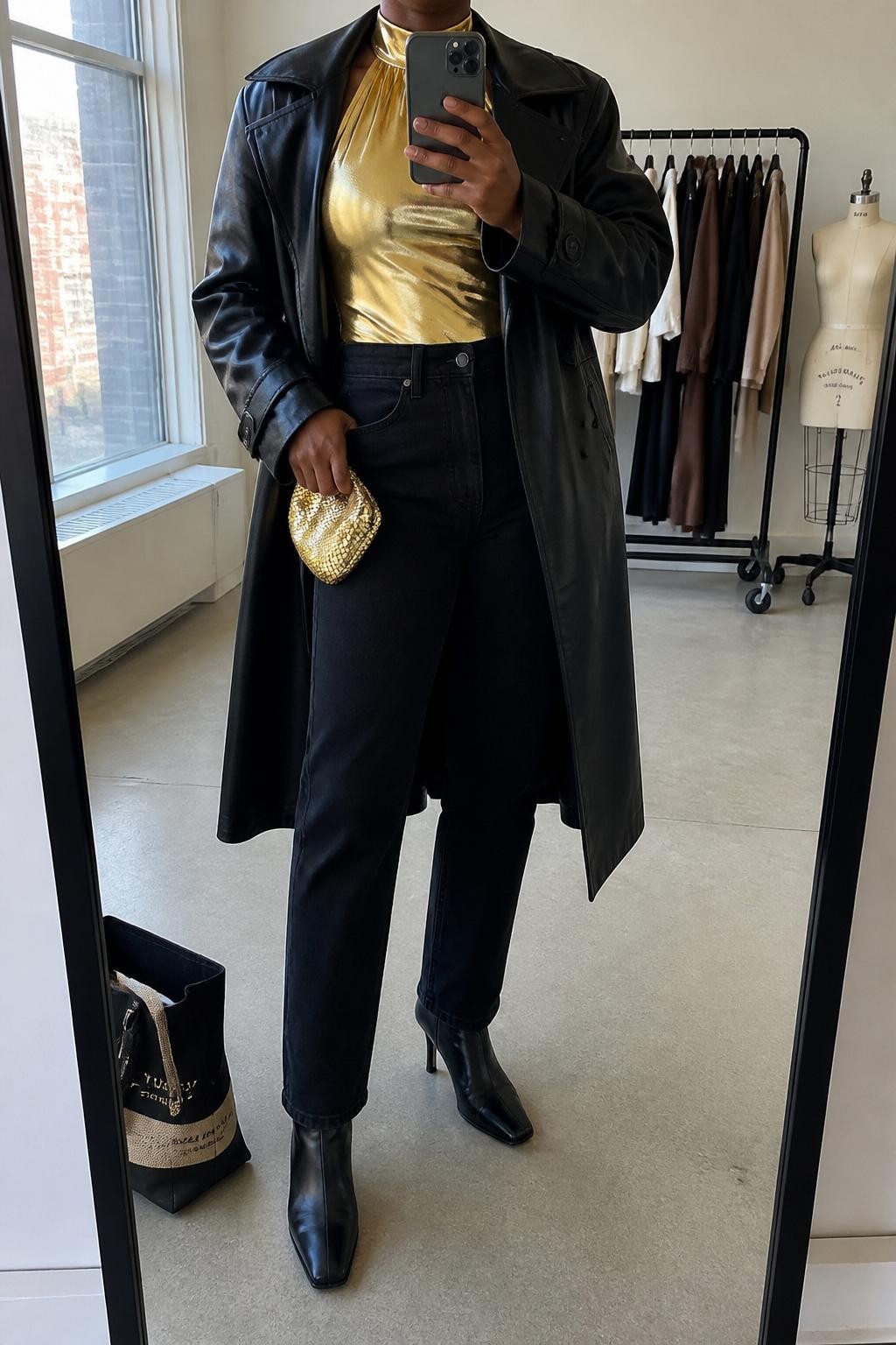 mirror selfie of a Black woman wearing the exact outfit described above, clearly showing a gold metallic halter top, black high-waisted straight jeans, black heeled ankle boots, black faux leather trench coat, and gold mini clutch, holding a phone in front of a mirror, cropped at NECK so the face does not show, with a confident casual selfie pose, in a fashion studio mirror corner, natural window light, including a tote bag resting on the floor and a clothing rack with garments behind, latest iPhone photo quality.