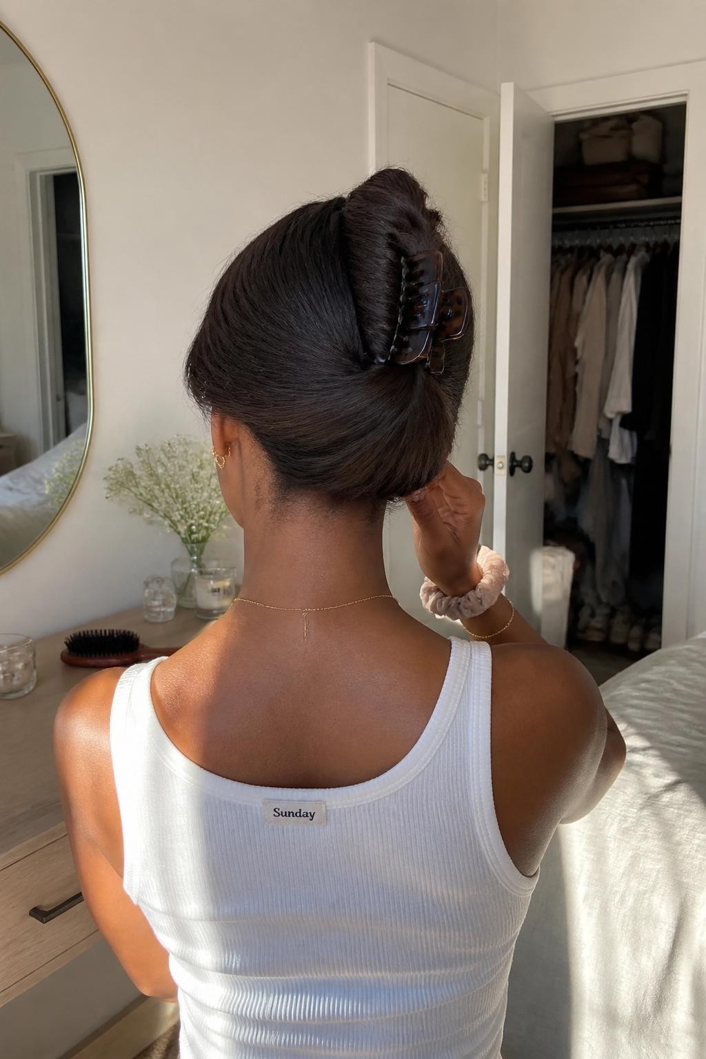 Image prompt: Photo of a Black woman wearing a soft French roll secured with a large tortoiseshell claw clip, back-of-head view to highlight the twist, sun-drenched bedroom with a slightly open closet behind her, natural window light, containing a hairbrush on the dresser and a silk scrunchie around her wrist, realistic quick getting-ready moment, latest iPhone photo quality.