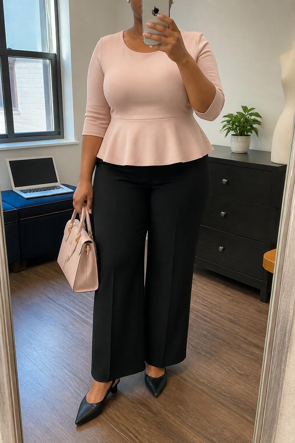 Mirror selfie of a plus size woman wearing black high-waisted wide-leg tailored trousers, a blush pink peplum top with three-quarter sleeves and a modest scoop neckline, black pointed slingback flats, a structured blush handbag, and pearl stud earrings, clearly showing the exact clothing items, black tailoring, blush peplum shape, footwear, and accessories listed, holding a phone in front of a mirror, cropped at the neck so the face is hidden, relaxed confident selfie pose, in a fashion studio mirror corner with natural window light, a laptop resting on a bench and a small plant on a dresser, realistic church outfit, latest iPhone photo quality.