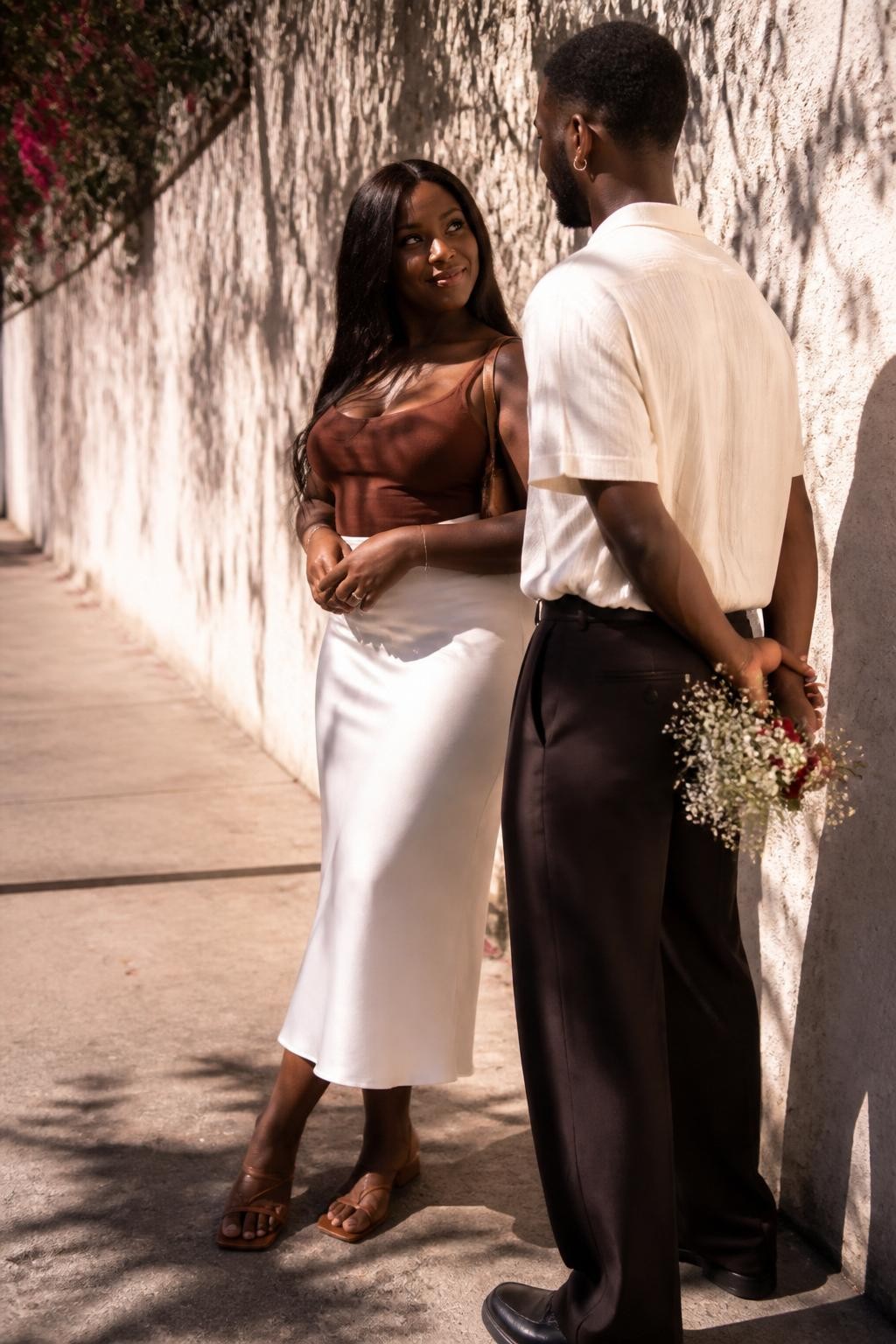 Image prompt: A beautiful DSLR couple photo on a sunny sidewalk beside a wall covered with flower shadows, featuring an adult Black woman with rich dark skin wearing a white slip skirt, fitted brown top, and strappy sandals, standing close to her partner as he gently holds a small bouquet behind his back, she is turned slightly toward him with a knowing smile, both bodies partly covered by soft leaf and flower shadows, warm afternoon light, textured cream wall, city sidewalk, realistic skin texture, natural clothing folds, editorial street couple photography, sweet and modern.