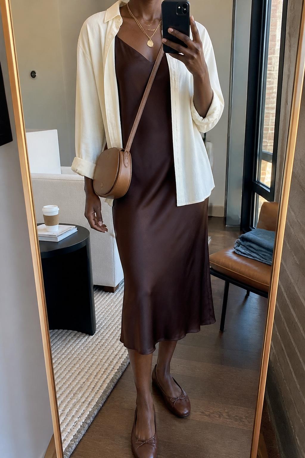 mirror selfie of a Black woman wearing the exact outfit described above, clearly showing a chocolate brown satin midi slip dress, lightweight cream button-down shirt worn open, brown leather ballet flats, a tan mini crossbody bag, and gold layered necklaces, holding a phone in front of a mirror, cropped at NECK so the face does not show, with a relaxed casual selfie pose, in a stylish city apartment, soft morning light, with a coffee cup on a side table and folded denim jeans on a chair, latest iPhone photo quality.
