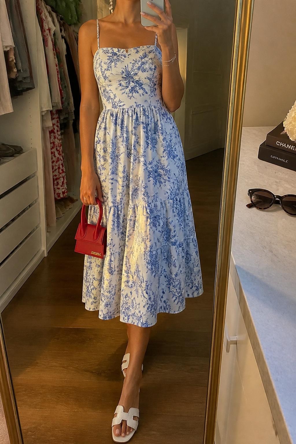 mirror selfie of a woman wearing a blue and white floral cotton sundress with thin straps and a flowing midi skirt, white flat sandals, red mini handbag, pearl drop earrings, and a slim silver bracelet, clearly showing the same clothing items, colors, fabrics, and accessories listed in the Outfit Pieces section, holding a phone in front of a mirror, cropped at NECK so the face does not show, with a relaxed casual selfie pose, in a walk-in closet mirror, evening golden hour, including an open closet with clothes visible and sunglasses placed on a dresser, latest iPhone photo quality.