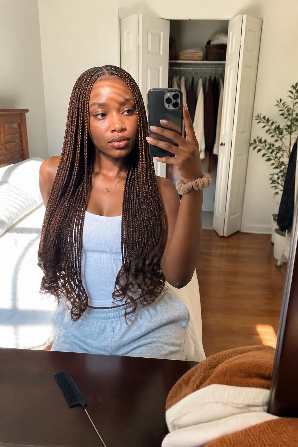 Image prompt: Photo of a Black woman wearing long chestnut knotless braids with soft curled tips and a neat middle part, candid mirror selfie, sun-drenched bedroom with a slightly open closet and folded fall sweaters behind her, natural window light, containing a rat-tail comb on the dresser and a silk scrunchie around her wrist, realistic everyday getting-ready moment, latest iPhone photo quality.