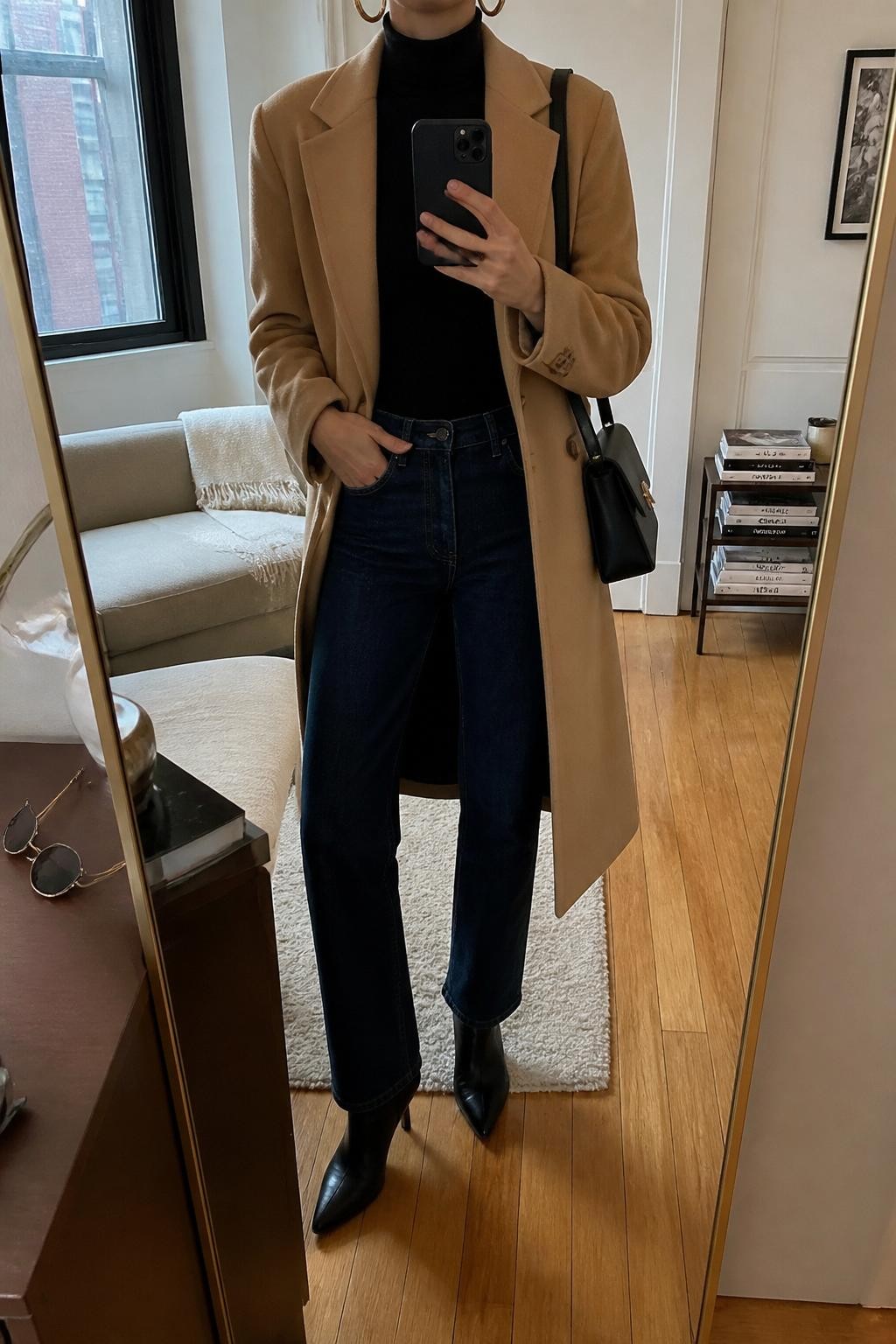 mirror selfie of a woman wearing the exact outfit described above, clearly showing a camel wool coat layered over a fitted black turtleneck, dark blue straight-leg jeans, black pointed ankle boots, a structured black shoulder bag, and gold hoop earrings, holding a phone in front of a mirror, cropped at NECK so the face does not show, with a relaxed casual selfie pose, in a stylish city apartment, natural window light, with a stack of fashion magazines on a side table and sunglasses placed on a dresser, latest iPhone photo quality.