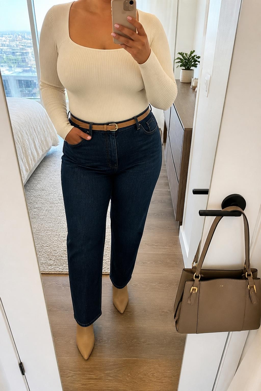 Mirror selfie of a plus size woman wearing dark indigo high-waisted straight-leg jeans, a fitted cream ribbed square-neck bodysuit, a slim camel leather belt, and pointed nude ankle boots, clearly showing the exact clothing items, colors, fabrics, and accessories listed, holding a phone in front of a mirror, cropped at the neck so her face does not show, relaxed casual selfie pose, in a stylish city apartment with natural window light, a structured handbag hanging on a door handle and a small plant on a dresser, natural unstaged appearance, latest iPhone photo quality.