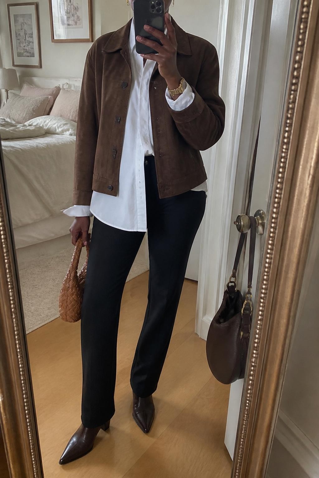 Mirror selfie of a Black woman wearing a chocolate brown suede jacket over an oversized white poplin button-down shirt and black slim straight-leg trousers, clearly showing dark brown pointed ankle boots, a woven tan handbag, and a slim gold wristwatch, holding an iPhone in front of a feminine-themed bedroom mirror, cropped at the neck so the face does not show, relaxed casual selfie pose, natural window light, soft pillows on an unmade bed and a handbag hanging on a door handle, natural unstaged setting, latest iPhone photo quality.