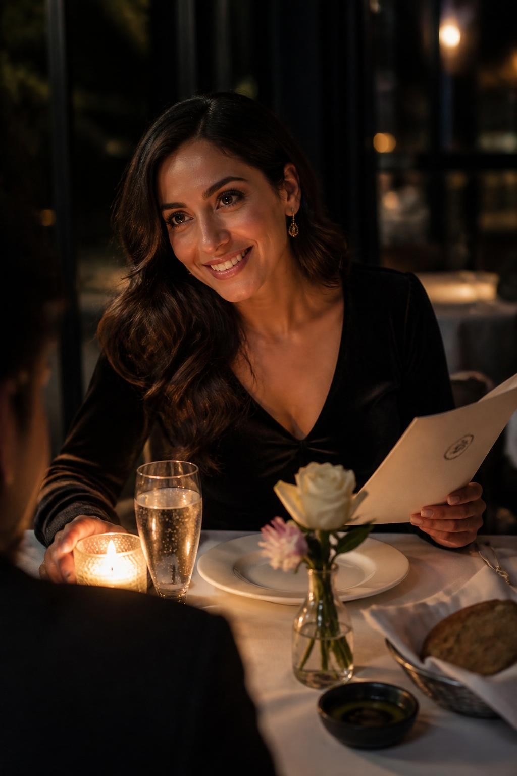 Image prompt: A beautiful DSLR restaurant portrait of an adult woman with medium skin tone leaning slightly across a small candlelit table, wearing a black velvet top, soft waves, and simple earrings, one hand resting near a candle, the other hand holding a folded menu, eyes looking toward someone across the table with a warm smile, white plates, sparkling water, bread plate, small flower vase, dark restaurant background, realistic candle glow on her face, soft shadows, natural expression, tasteful dinner photography with an intimate but relaxed mood.
