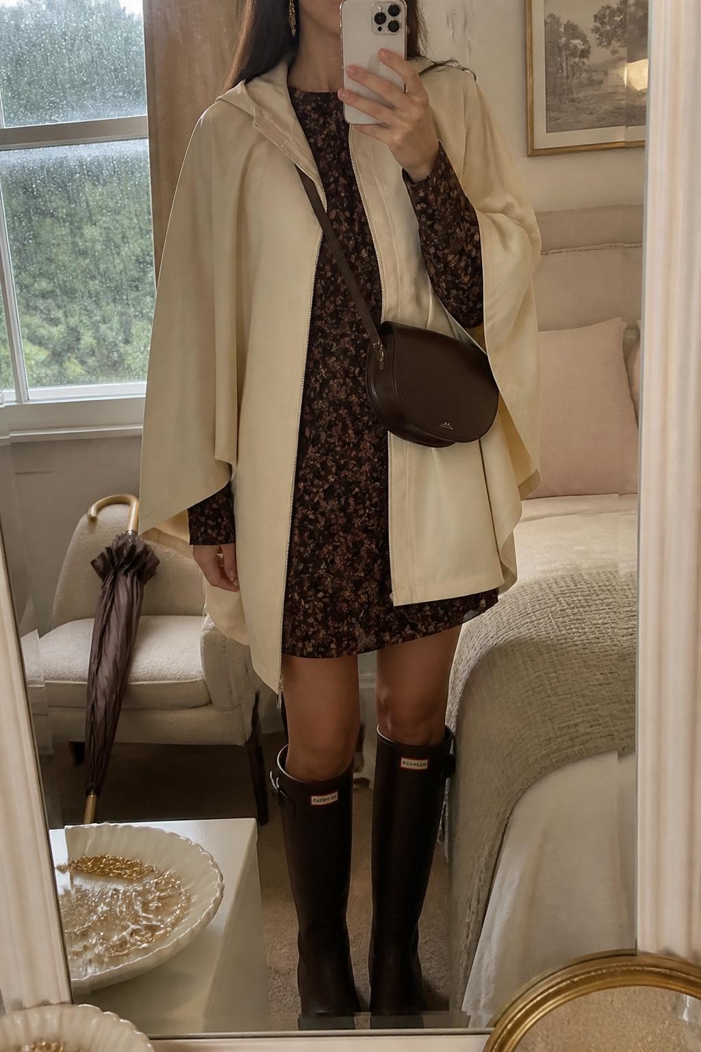 mirror selfie of a woman wearing the exact outfit described above, clearly showing a cream water-resistant rain cape layered over a dark floral long-sleeve mini dress with burgundy and brown flowers, dark brown knee-high rain boots, a chocolate crossbody bag, and gold drop earrings, holding a phone in front of a mirror, cropped at NECK so the face does not show, with a relaxed casual selfie pose, in a feminine bedroom mirror setup, soft gray rainy window light, with a jewelry tray on the vanity and a folded umbrella on a chair, latest iPhone photo quality.