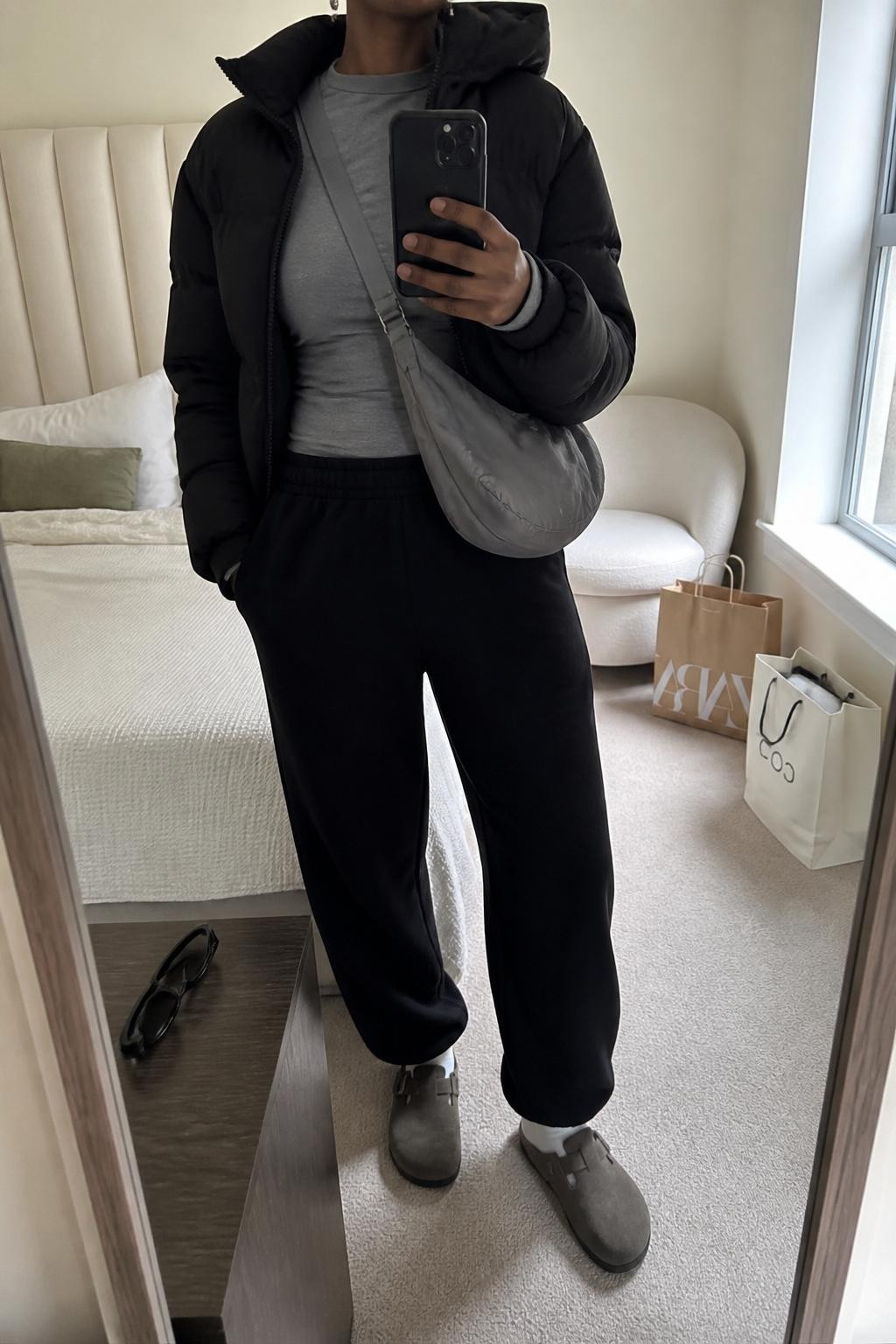 mirror selfie of a Black woman wearing the exact outfit described above, clearly showing dark taupe suede Birkenstock-style clogs with white socks, black wide-leg sweatpants, a fitted gray long-sleeve top, a cropped black puffer jacket, a gray nylon crossbody bag, and silver stud earrings, holding a phone in front of a mirror, cropped at NECK so the face does not show, with a relaxed casual selfie pose, in a cozy bedroom mirror setup, natural window light, with shopping bags beside a chair and sunglasses placed on a dresser, latest iPhone photo quality.