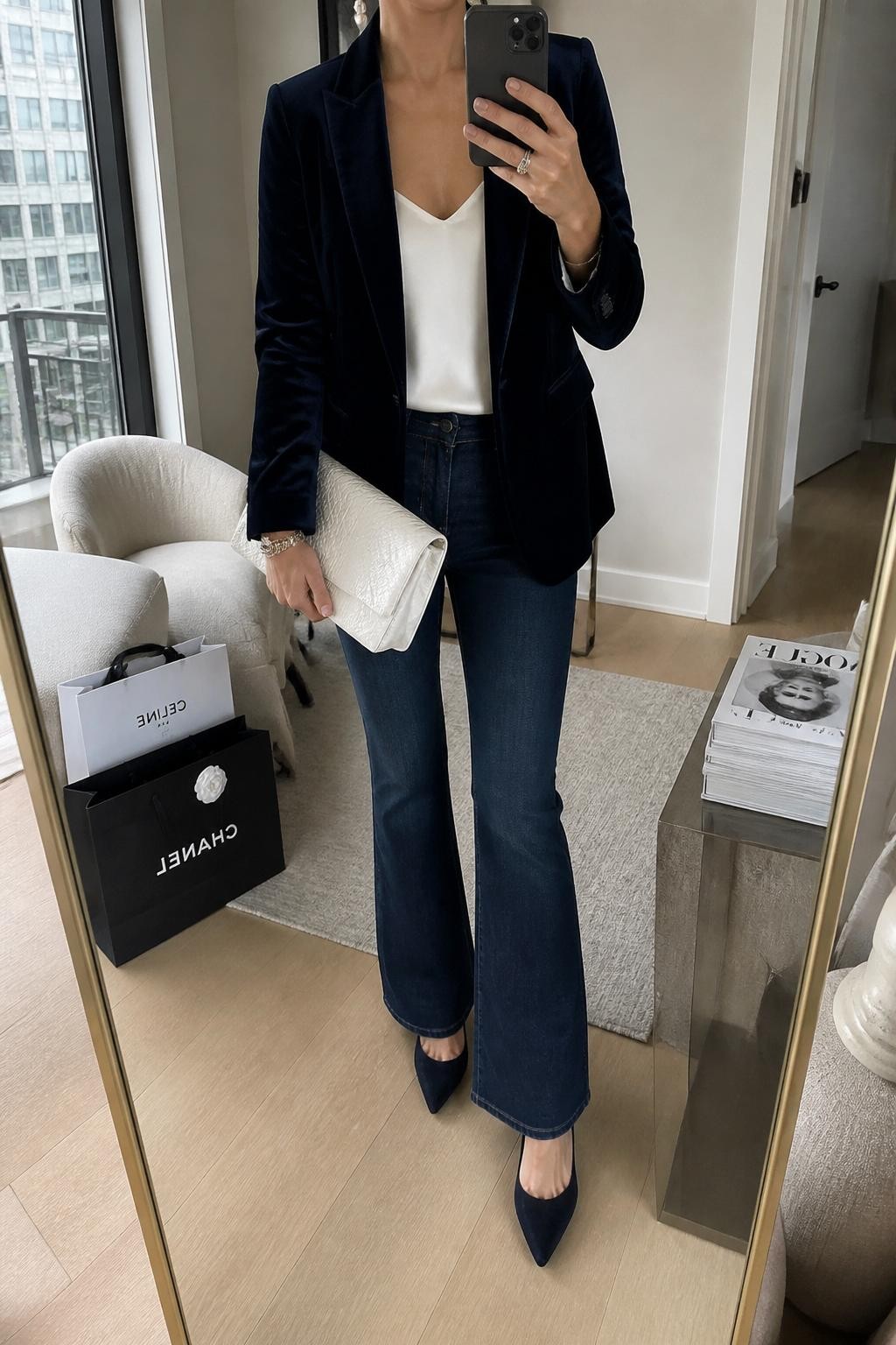 mirror selfie of a woman wearing the exact outfit described above, clearly showing a navy velvet blazer layered over an ivory satin cami, dark wash flare jeans, navy pointed pumps, a pearl white clutch, and silver earrings, holding a phone in front of a mirror, cropped at NECK so the face does not show, with a relaxed casual selfie pose, in a stylish city apartment, natural window light, with shopping bags beside a chair and a stack of fashion magazines on a side table, latest iPhone photo quality.
