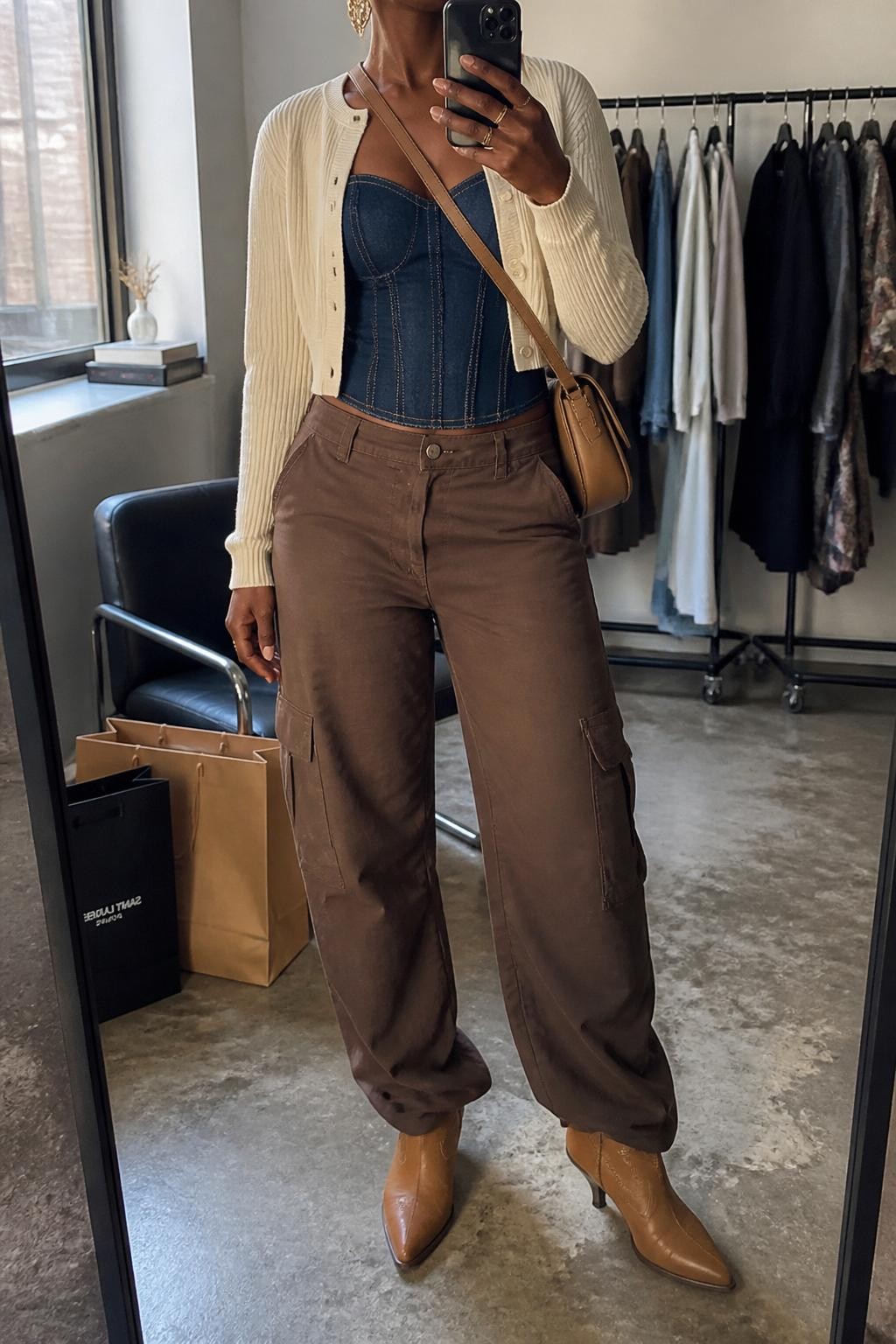 mirror selfie of a Black woman wearing the exact outfit described above, clearly showing a structured dark denim corset top, brown relaxed cargo pants, tan western ankle boots, a cropped cream cardigan, a tan crossbody bag, and gold statement earrings, holding a phone in front of a mirror, cropped at NECK so the face does not show, with a relaxed casual selfie pose, in a fashion studio mirror corner, natural window light, with clothing rack with garments behind and shopping bags beside a chair, latest iPhone photo quality.