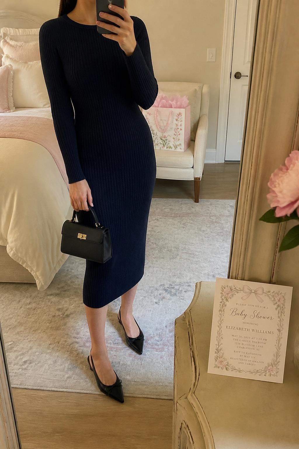 mirror selfie of a woman wearing the exact outfit described above, clearly showing a navy long-sleeve knit midi dress, black slingback flats, a pearl headband, a small black top-handle bag, and pearl stud earrings, holding a phone in front of a mirror, cropped at NECK so the face does not show, with a relaxed casual selfie pose, in a feminine bedroom mirror setup, warm natural light, with a baby shower invitation on the dresser and a pastel gift bag on a chair, latest iPhone photo quality.