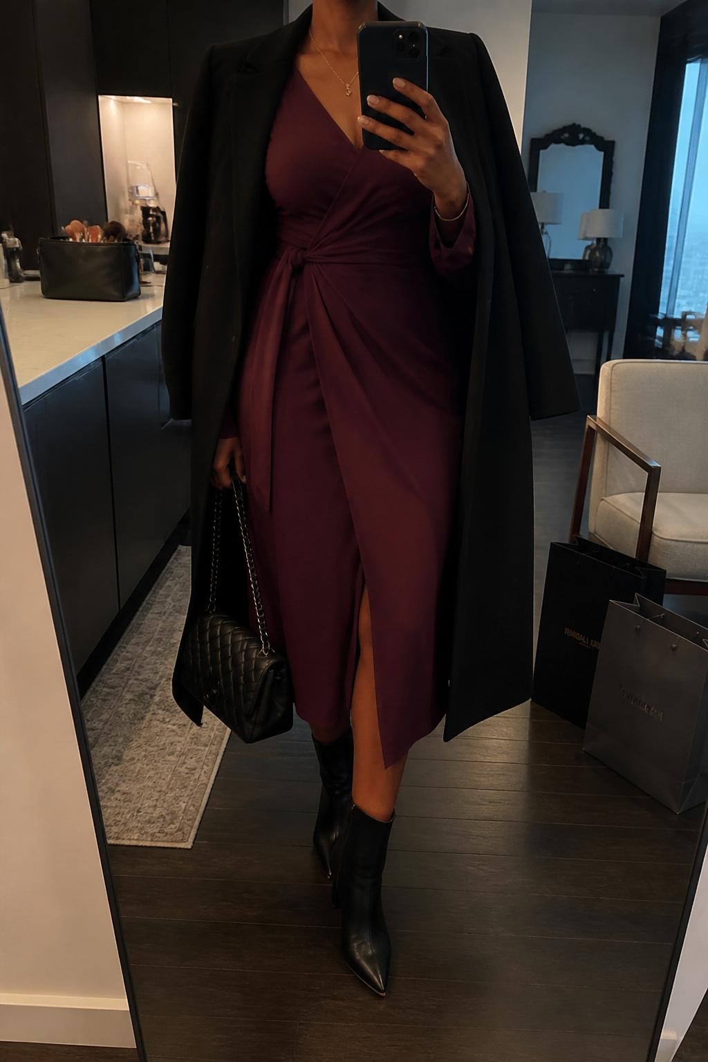 mirror selfie of a Black woman wearing the exact outfit described above, clearly showing a plum wrap midi dress with long sleeves, black wool coat draped open, black pointed ankle boots, a black quilted handbag, and a delicate gold necklace, holding a phone in front of a mirror, cropped at NECK so the face does not show, with a relaxed casual selfie pose, in a stylish city apartment, warm moody lighting, with a makeup brush or cosmetic bag on the counter and shopping bags beside a chair, latest iPhone photo quality.