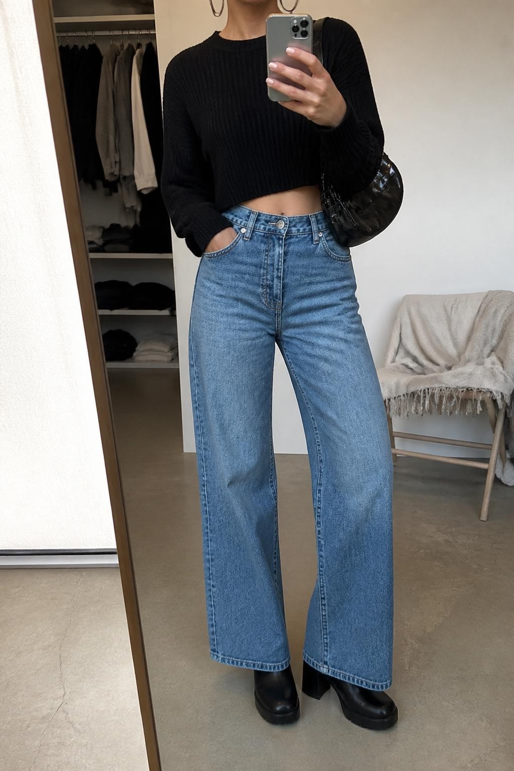 Mirror selfie of a woman wearing a black cropped knit sweater, high-waisted medium-wash wide-leg jeans, black platform ankle boots, silver hoop earrings, and a black leather shoulder bag, holding a phone in front of a mirror, cropped at NECK so the face does not show, relaxed casual selfie pose, in a minimal dressing room, soft indoor lighting, with an open closet with clothes visible and folded blanket on a chair, latest iPhone photo quality.