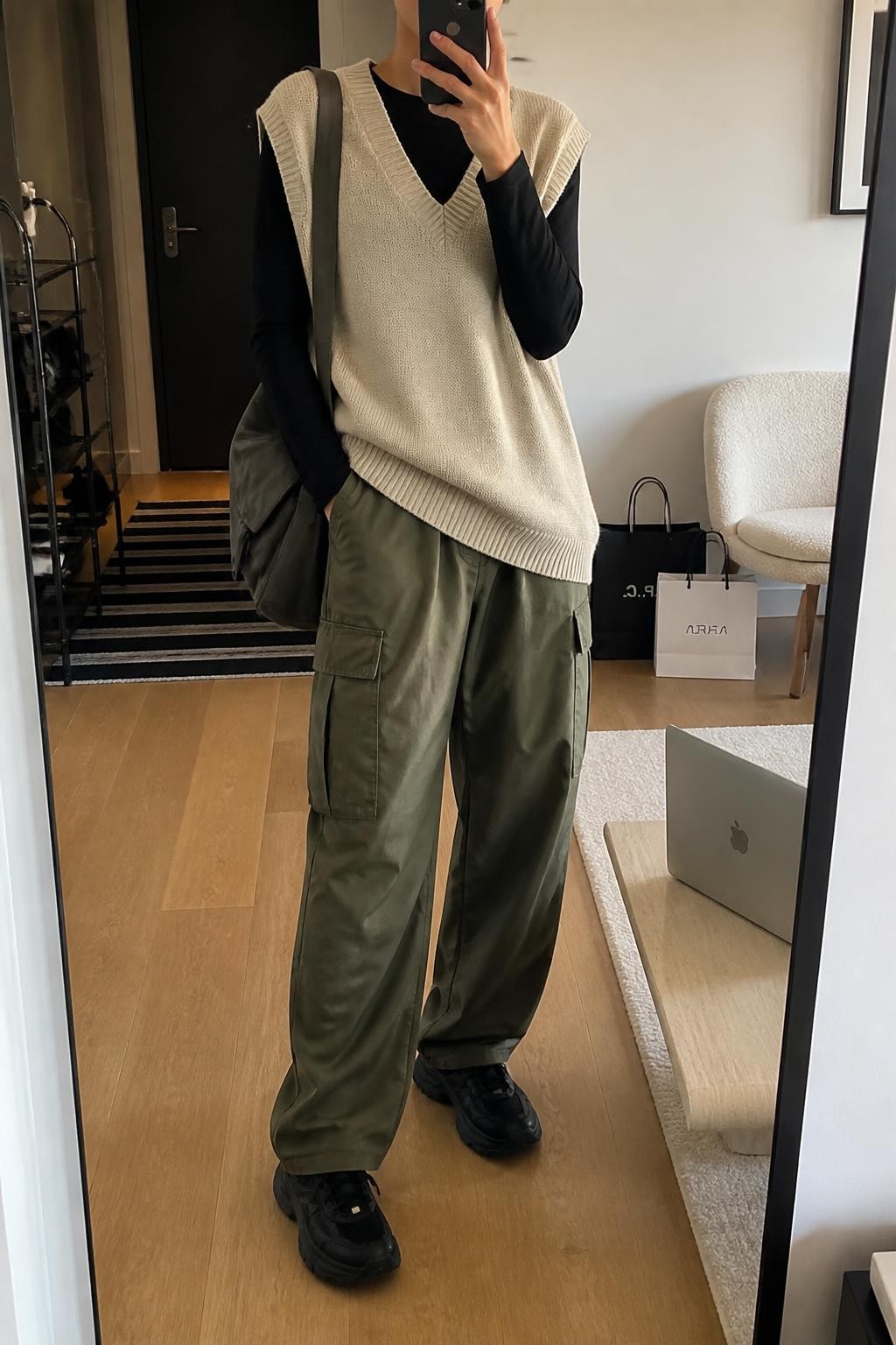Mirror selfie of a woman wearing an oversized oatmeal V-neck sweater vest over a fitted black long-sleeve top, loose olive-green cargo pants, chunky black leather sneakers, and a charcoal-grey messenger bag, clearly showing the exact clothing items, colors, fabrics, and accessories listed, holding a phone in front of a stylish city apartment mirror, cropped at the neck so the face does not show, relaxed casual selfie pose, soft indoor lighting, a laptop on a bench and shopping bags beside a chair, modern realistic setting, latest iPhone photo quality.
