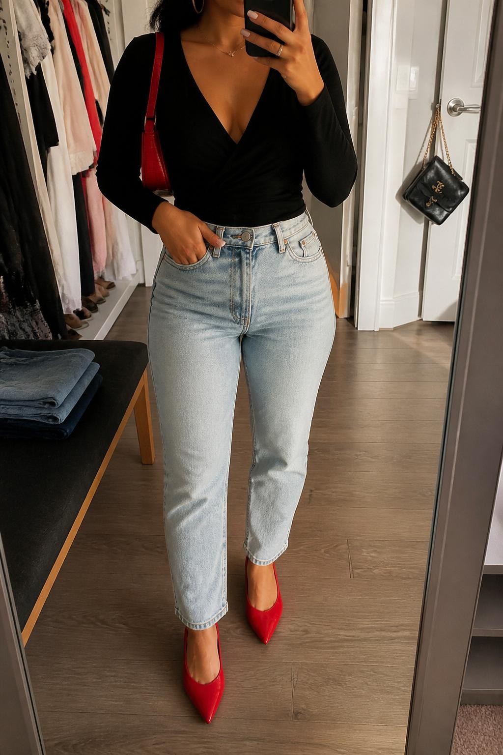 Mirror selfie of a Latina woman wearing a black wrap top, light wash high-waisted mom jeans, red pointed flats, gold hoop earrings, and a small red shoulder bag, holding a phone in front of a mirror, cropped at NECK so the face does not show, relaxed casual selfie pose, in a walk-in closet mirror, evening golden hour, with folded denim jeans on a bench and a handbag hanging on a door handle, latest iPhone photo quality.