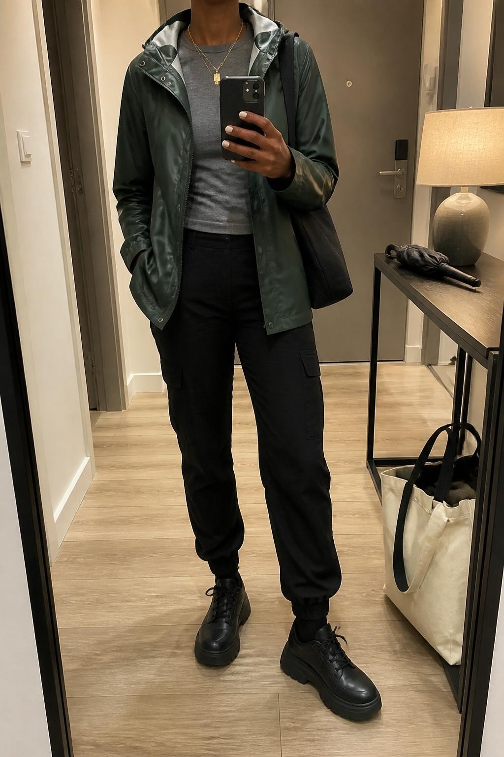 mirror selfie of a Black woman wearing the exact outfit described above, clearly showing a forest green slicker jacket layered over a fitted gray long-sleeve top, black tapered cargo pants, black waterproof platform sneakers, a black nylon shoulder bag, and gold layered necklaces, holding a phone in front of a mirror, cropped at NECK so the face does not show, with a relaxed casual selfie pose, in a modern apartment hallway mirror, warm indoor lighting, with a folded umbrella on a console table and a tote bag on the floor, latest iPhone photo quality.