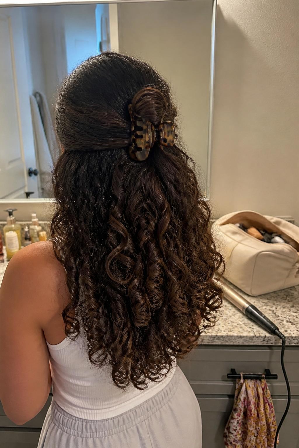 Image prompt: Photo of a woman wearing long natural curls styled in a half-up twist secured with a large tortoiseshell claw clip, back-of-head view to highlight the details, bathroom vanity mirror with a makeup bag nearby, soft indoor lighting, containing a curling iron on the counter and a silk scarf hanging from a drawer, realistic everyday styling moment, latest iPhone photo quality.