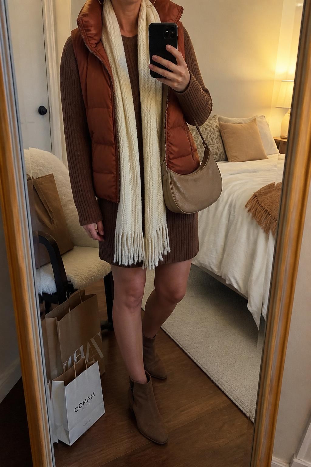 Mirror selfie of a woman wearing a brown ribbed sweater dress, rust quilted puffer vest, cream knit scarf, brown suede ankle boots, gold hoop earrings, and a tan crescent bag, holding a phone in front of a mirror, cropped at NECK so the face does not show, relaxed casual selfie pose, in a cozy bedroom mirror setup, evening golden hour, with shopping bags beside a chair and soft pillows on an unmade bed, latest iPhone photo quality.