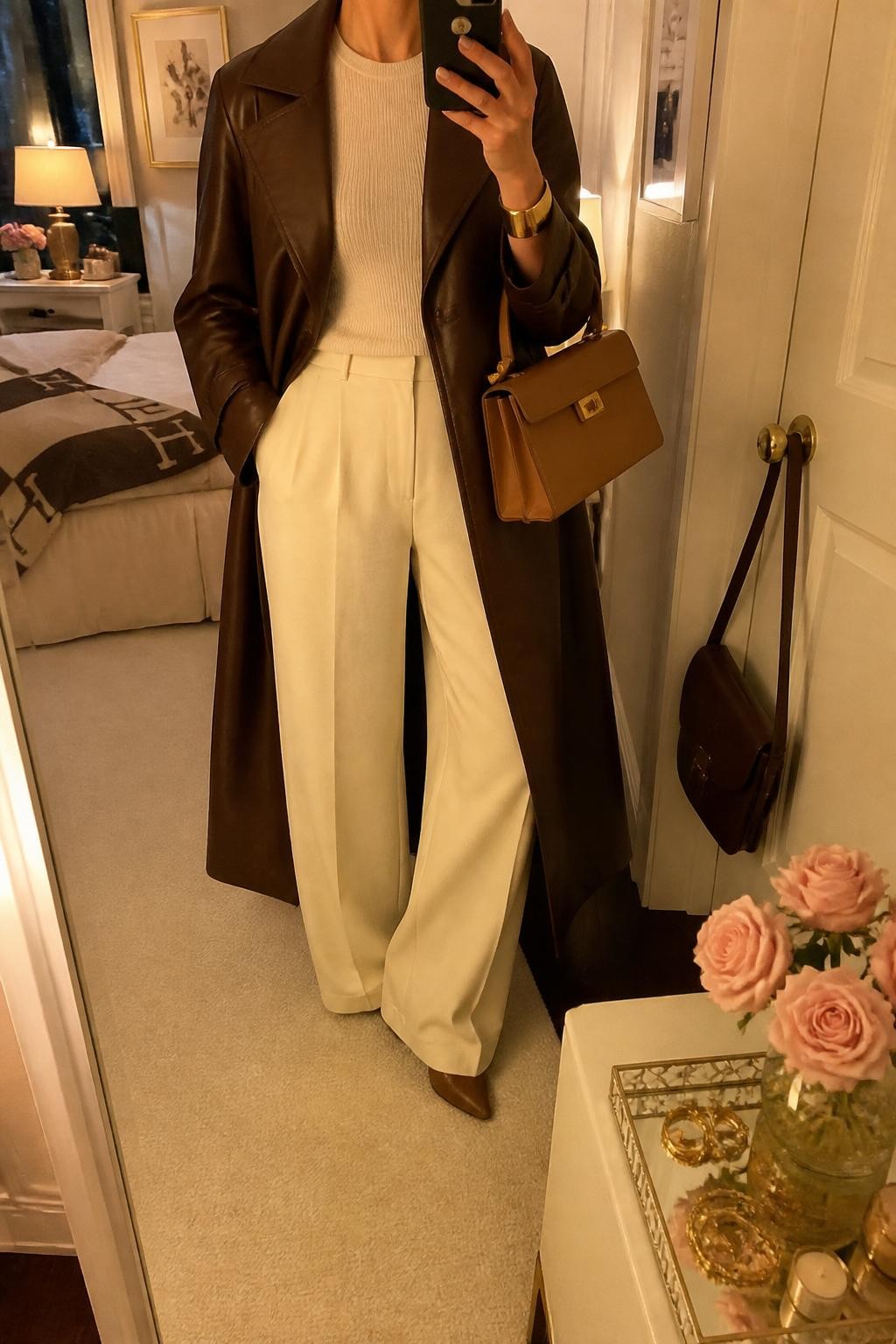 mirror selfie of a woman wearing the exact outfit described above, clearly showing an espresso brown leather trench coat layered over a fitted oatmeal knit top, cream wide-leg trousers, brown pointed boots, a tan top-handle bag, and gold cuff bracelet, holding a phone in front of a mirror, cropped at NECK so the face does not show, with a relaxed casual selfie pose, in a feminine-themed bedroom, evening golden hour, with a handbag hanging on a door handle and a jewelry tray on the vanity, latest iPhone photo quality.