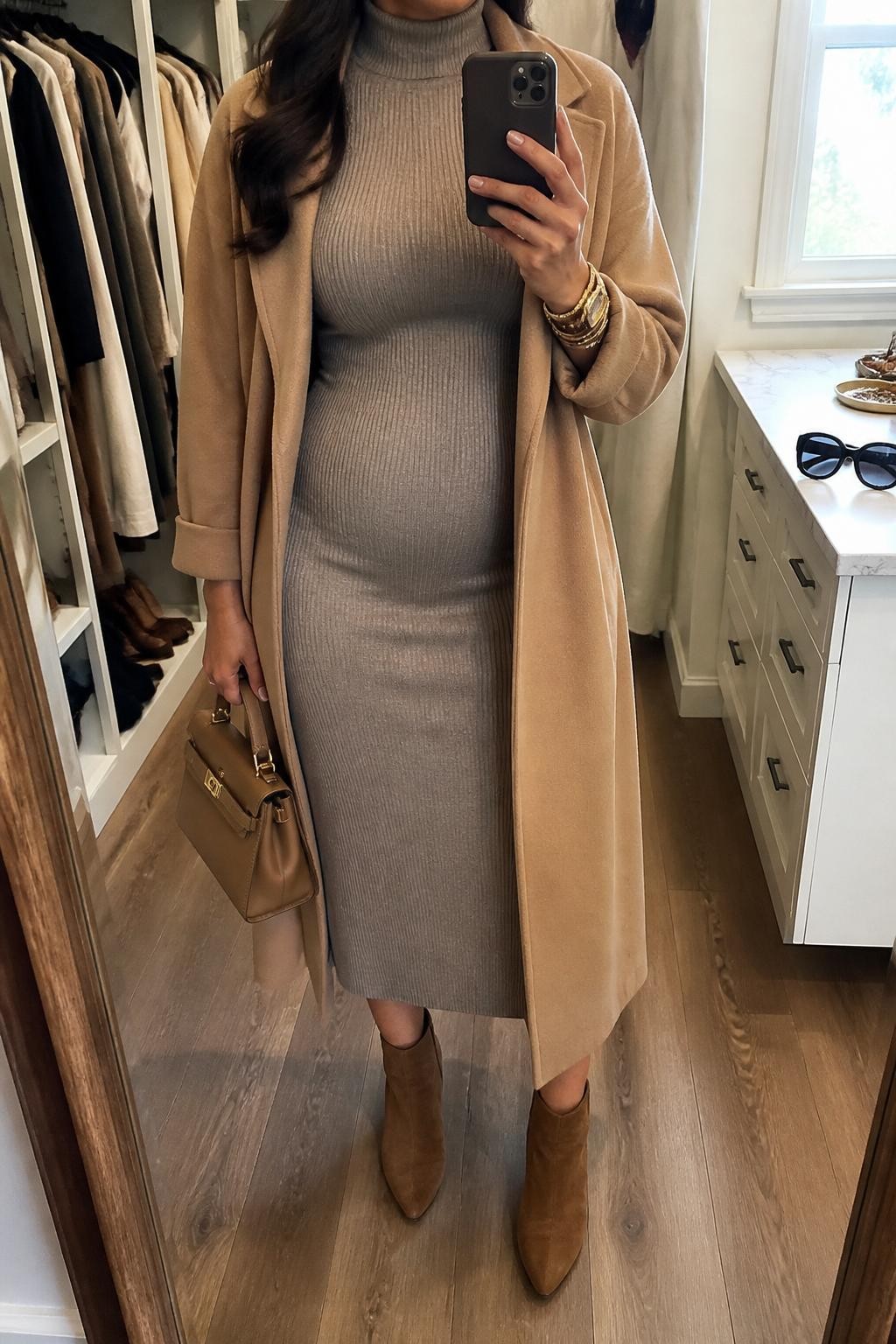 Mirror selfie of a curvy woman wearing a taupe turtleneck sweater midi dress, long camel coat, brown suede ankle boots, gold bracelet stack, and a tan top-handle bag, holding a phone in front of a mirror, cropped at NECK so the face does not show, relaxed casual selfie pose, in a walk-in closet mirror, natural window light, with an open closet with clothes visible and sunglasses placed on a dresser, latest iPhone photo quality.