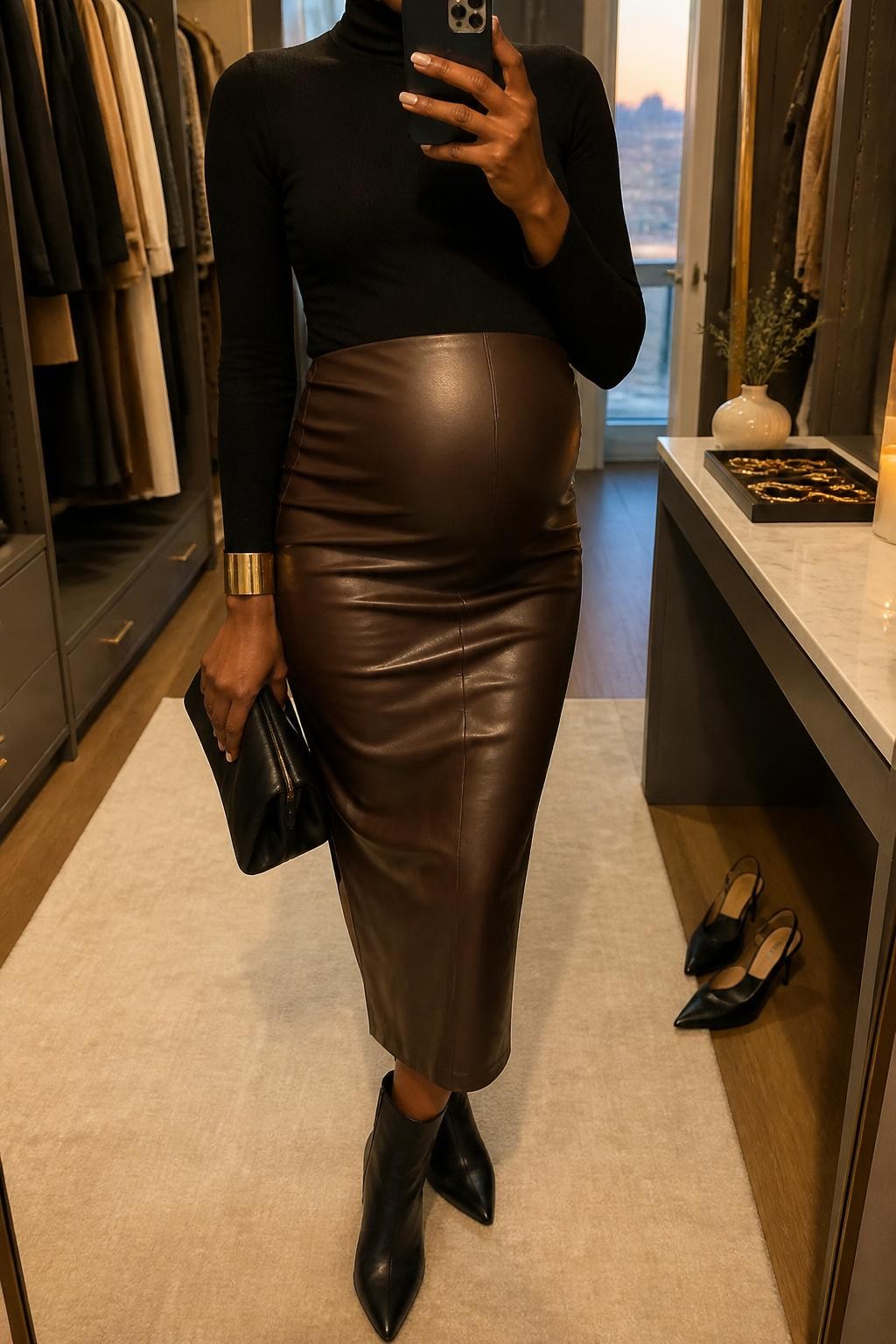 mirror selfie of a pregnant Black woman wearing a fitted black maternity turtleneck, dark brown faux leather maternity midi skirt, black pointed ankle boots, a gold cuff bracelet, and a black clutch bag, clearly showing the same clothing items, colors, fabrics, and accessories listed in the Outfit Pieces section, holding a phone in front of a mirror, cropped at NECK so the face does not show, with a relaxed casual selfie pose, in a walk-in closet mirror, evening golden hour, including a jewelry tray on the vanity and a pair of heels on the floor nearby, latest iPhone photo quality.