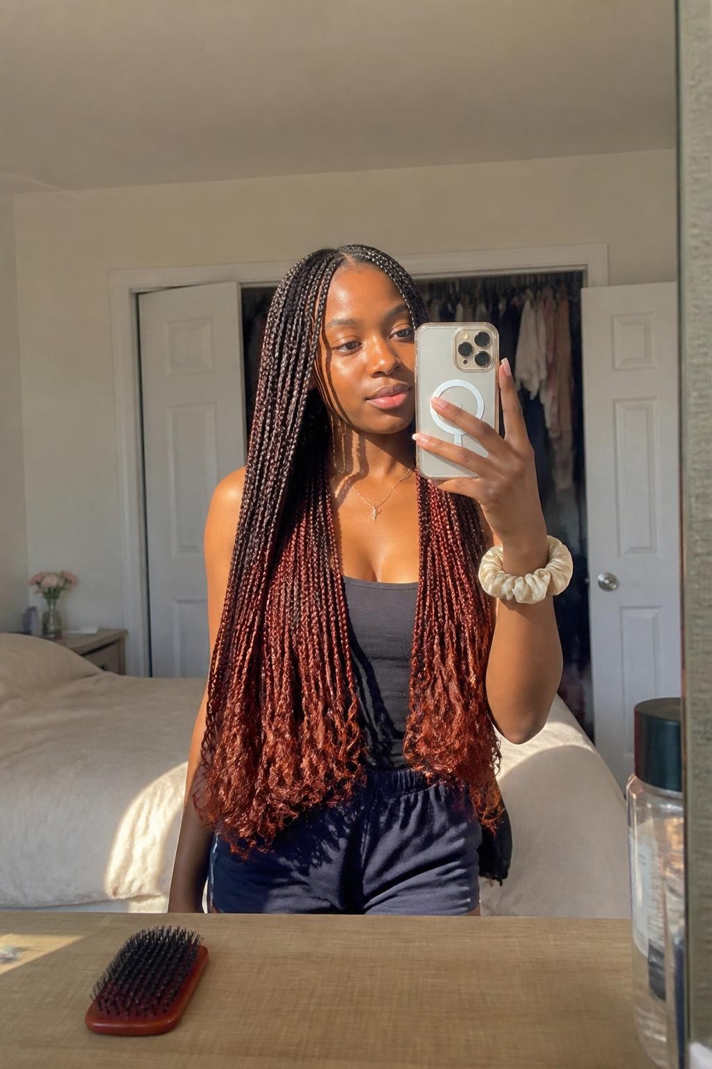 Image prompt: Photo of a Black woman wearing waist-length knotless braids with rich copper-colored tips and a neat middle part, candid mirror selfie, sun-drenched bedroom with a slightly open closet behind her, natural window light, containing a silk scrunchie on her wrist and a hairbrush resting on the dresser, realistic getting-ready moment, latest iPhone photo quality.