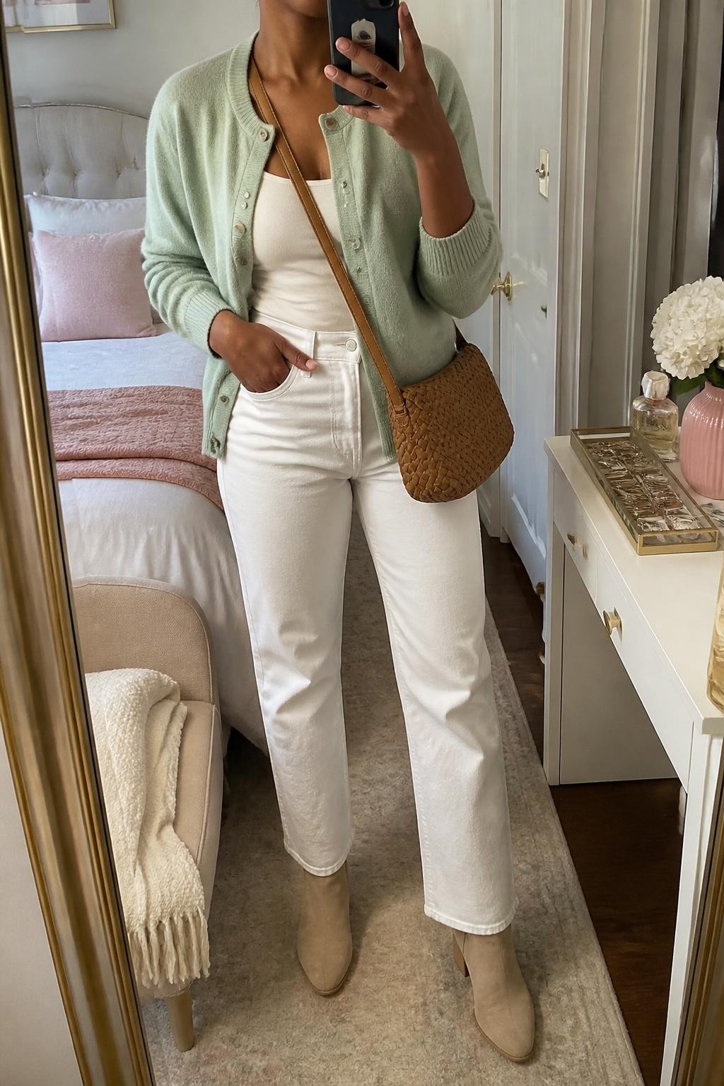 Mirror selfie of a Black woman wearing a soft seafoam-green button-front cardigan over a fitted ivory camisole, white high-waisted straight-leg jeans, beige suede ankle boots, and a woven tan leather crossbody bag, clearly showing the exact clothing items, colors, fabrics, and accessories listed, holding a phone in front of a feminine-themed bedroom mirror, cropped at the neck so the face does not show, relaxed casual selfie pose, soft indoor lighting, a folded blanket on a chair and a jewelry tray on the vanity, realistic lived-in setting, latest iPhone photo quality.