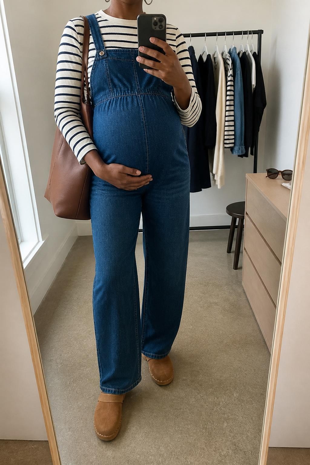 mirror selfie of a pregnant Black woman wearing a dark blue denim maternity jumpsuit over a cream and navy striped long sleeve tee, tan suede clogs, a brown leather tote bag, and small silver hoops, clearly showing the same clothing items, colors, fabrics, and accessories listed in the Outfit Pieces section, holding a phone in front of a mirror, cropped at NECK so the face does not show, with a relaxed casual selfie pose, in a minimal dressing room, natural window light, including a clothing rack with garments behind and sunglasses placed on a dresser, latest iPhone photo quality.