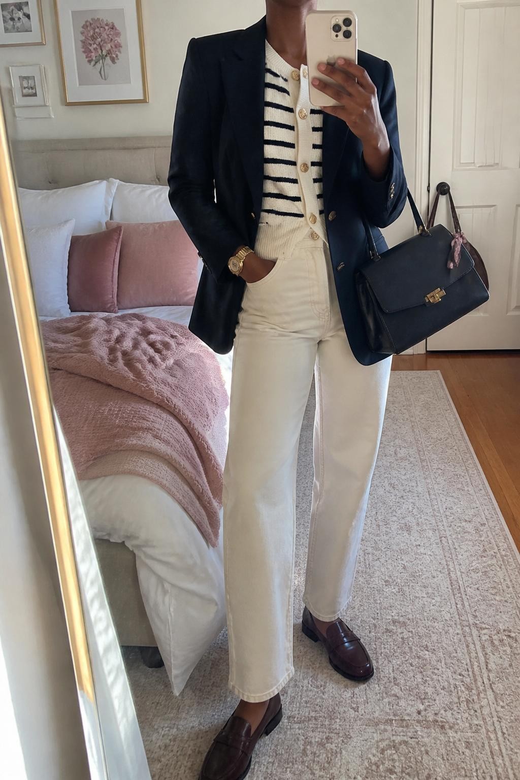 Mirror selfie of a Black woman wearing a navy tailored blazer over a cream-and-navy striped knit cardigan buttoned as a top, clearly showing ecru straight-leg jeans, oxblood leather loafers, a structured navy handbag, and a slim gold wristwatch, holding an iPhone in front of a feminine-themed bedroom mirror, cropped at the neck so the face does not show, relaxed casual selfie pose, natural window light, soft pillows on an unmade bed and a handbag hanging on a door handle, natural unstaged setting, latest iPhone photo quality.