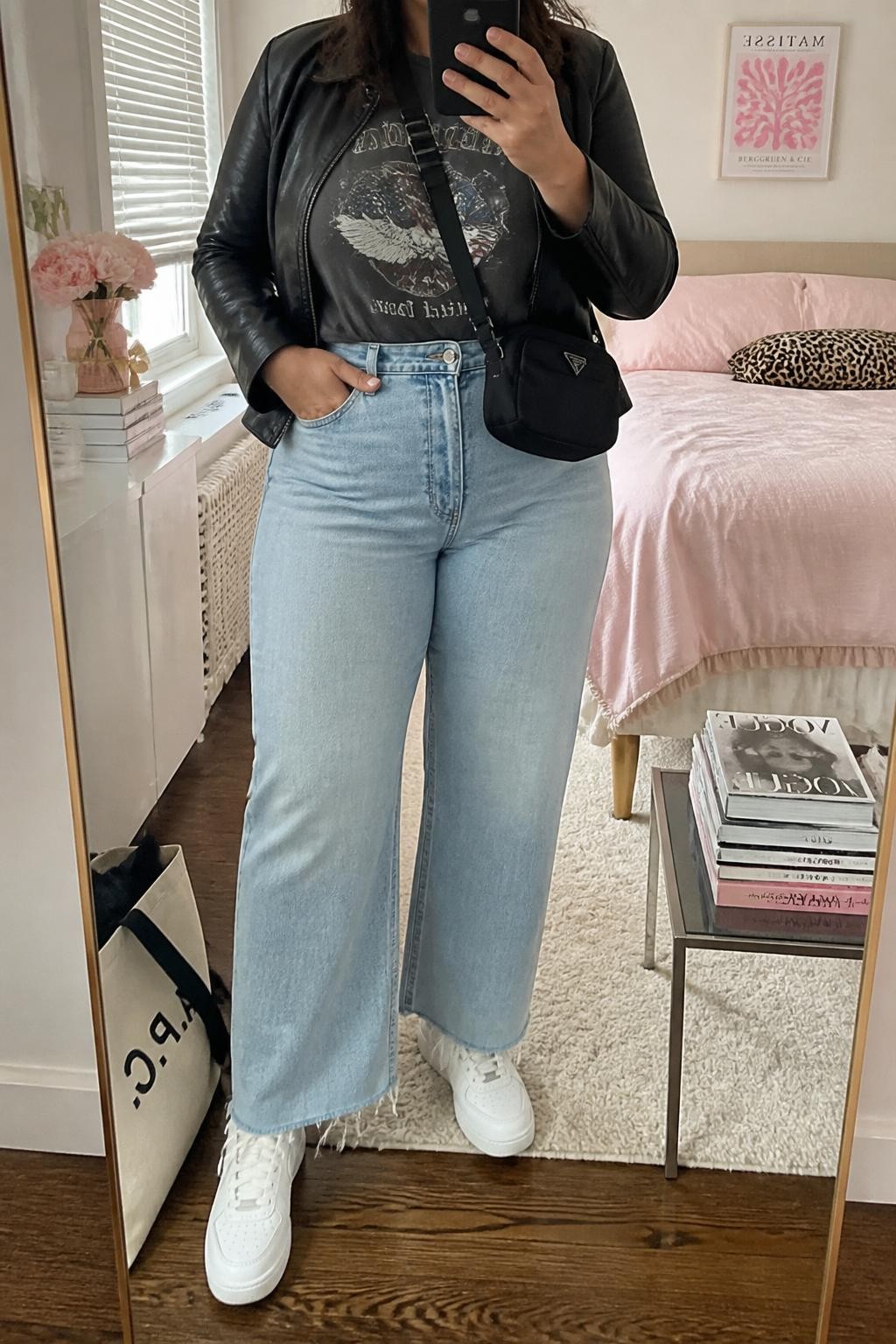 Mirror selfie of a plus size woman wearing light blue high-waisted wide-leg jeans with a raw ankle-length hem, a fitted faded charcoal graphic T-shirt tucked into the waistband, a cropped black leather jacket, white leather sneakers, and a small black nylon crossbody bag, clearly showing the exact clothing items, light denim, raw hem, graphic print, black jacket, footwear, and accessories listed, holding a phone in front of a mirror, cropped at the neck so her face does not show, relaxed casual selfie stance, in a feminine-themed bedroom with natural window light, a stack of fashion magazines on a side table and a tote bag resting on the floor, realistic weekend outfit, latest iPhone photo quality.