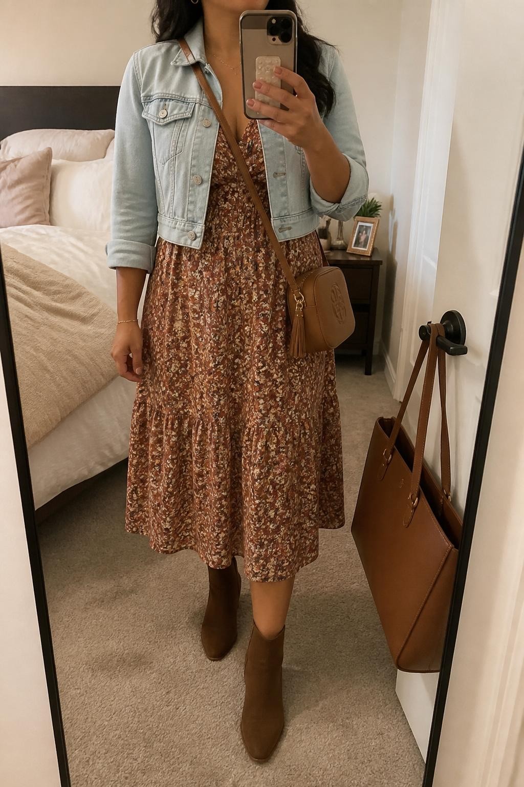 Mirror selfie of a Latina woman wearing a warm-toned floral midi dress, cropped light-wash denim jacket, brown ankle boots, small gold hoops, and a tan crossbody bag, holding a phone in front of a mirror, cropped at NECK so the face does not show, relaxed casual selfie pose, in a cozy bedroom mirror setup, soft indoor lighting, with soft pillows on an unmade bed and a handbag hanging on a door handle, latest iPhone photo quality.