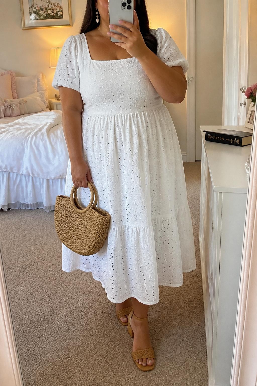 Mirror selfie of a plus size woman wearing a white cotton eyelet midi dress with short puff sleeves, a square neckline, a fitted waist, and a softly flared skirt, tan woven block-heel sandals, a natural woven handbag, and pearl drop earrings, clearly showing the exact dress, white eyelet texture, tan footwear, woven bag, and accessories listed, holding a phone in front of a mirror, cropped at the neck so her face is hidden, relaxed feminine selfie stance, in a feminine-themed bedroom with evening golden-hour lighting, soft pillows on an unmade bed and a Bible resting on a dresser, realistic warm-weather church outfit, latest iPhone photo quality.