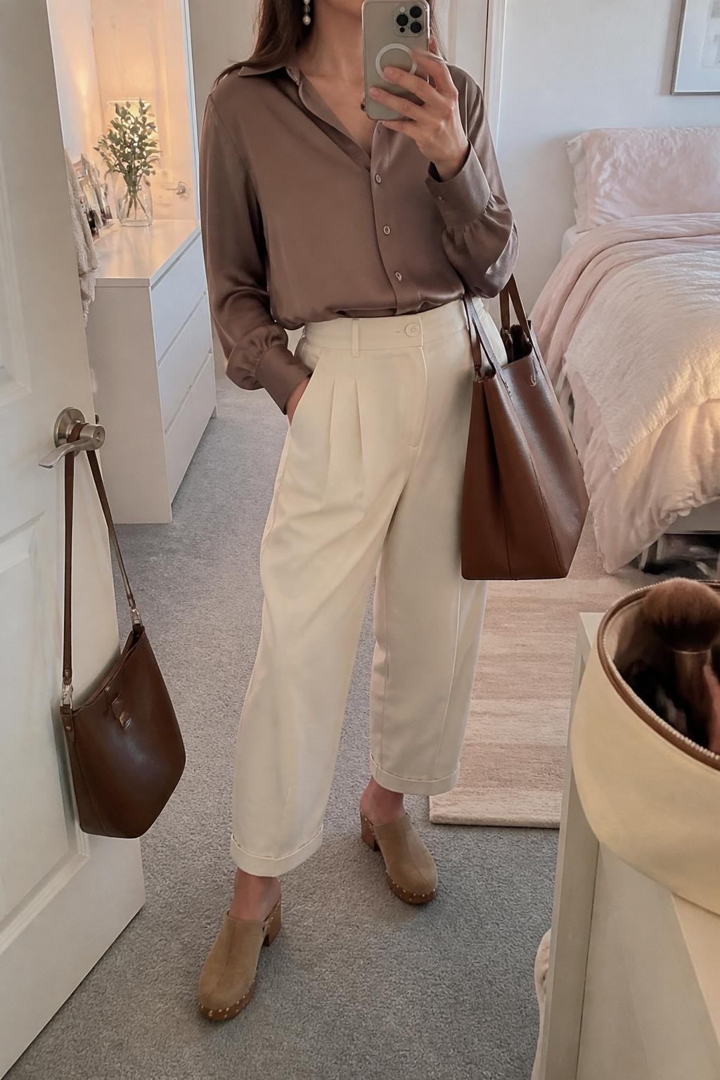 mirror selfie of a woman wearing the exact outfit described above, clearly showing a mocha satin blouse with long sleeves tucked into cream barrel pants, tan suede clogs, a brown structured tote bag, and pearl drop earrings, holding a phone in front of a mirror, cropped at NECK so the face does not show, with a relaxed casual selfie pose, in a feminine-themed bedroom, soft indoor lighting, with a makeup brush or cosmetic bag on the counter and a handbag hanging on a door handle, latest iPhone photo quality.