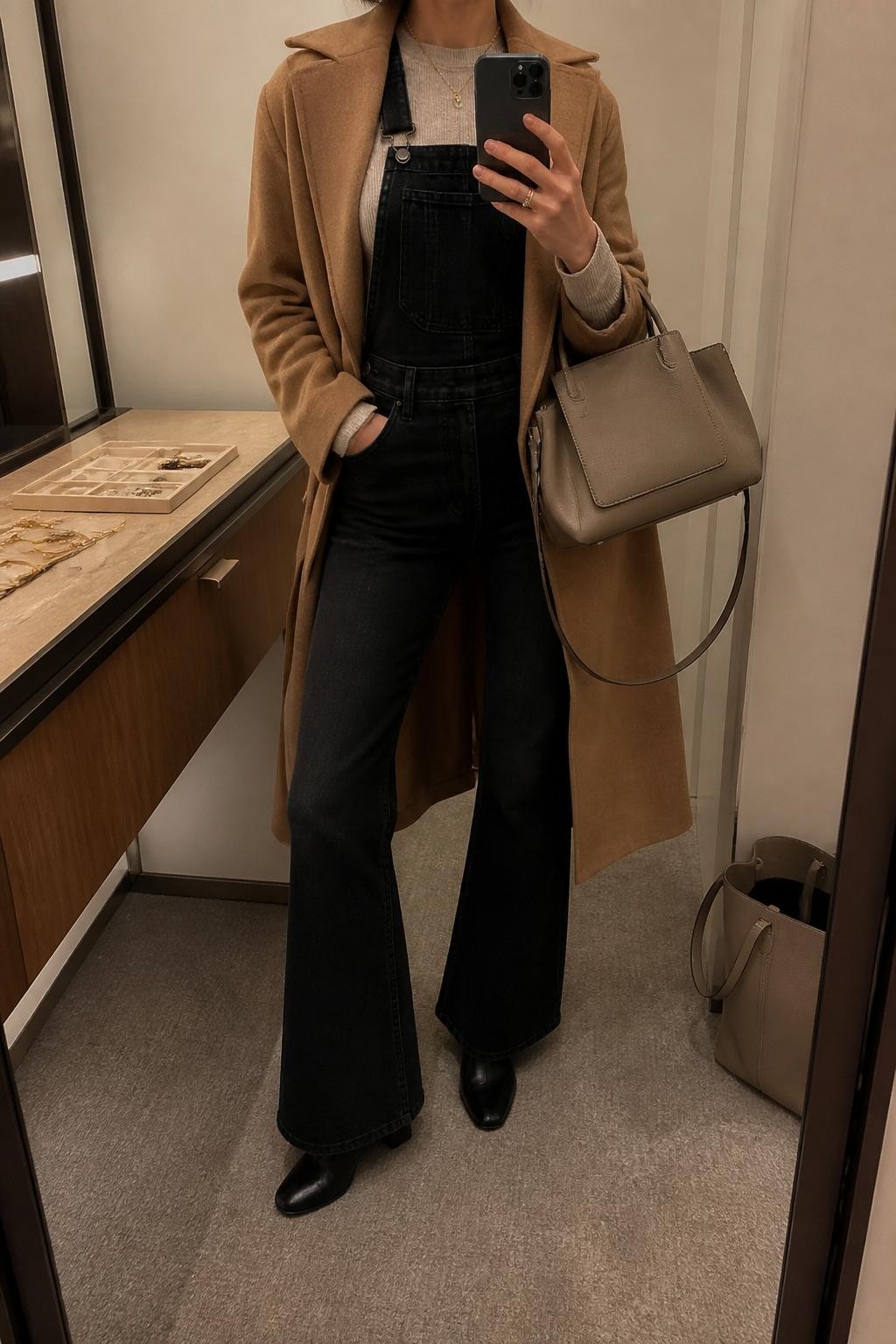 Mirror selfie of a woman wearing black denim flared overalls over a fitted oatmeal long-sleeve knit top with a camel wool coat layered on top, clearly showing black heeled ankle boots and a structured taupe handbag, holding an iPhone in front of a minimal dressing room mirror, cropped at the neck so the face does not show, relaxed casual selfie pose, warm moody lighting, a jewelry tray on the vanity and a tote bag resting on the floor, natural unstaged setting, latest iPhone photo quality.