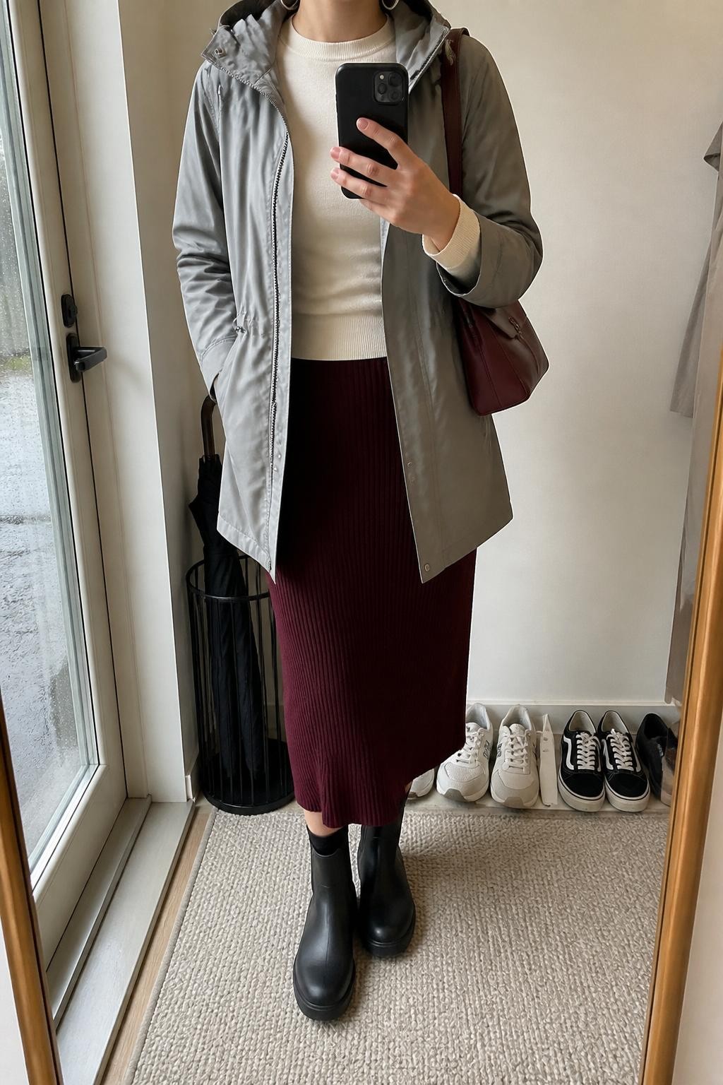 mirror selfie of a woman wearing the exact outfit described above, clearly showing a gray hooded water-resistant coat layered over a cream fitted sweater, burgundy ribbed knit midi skirt, black rubber ankle boots, a burgundy mini shoulder bag, and silver hoop earrings, holding a phone in front of a mirror, cropped at NECK so the face does not show, with a relaxed casual selfie pose, in a minimal entryway mirror, soft rainy daylight, with shoes lined neatly by the door and a small umbrella stand nearby, latest iPhone photo quality.