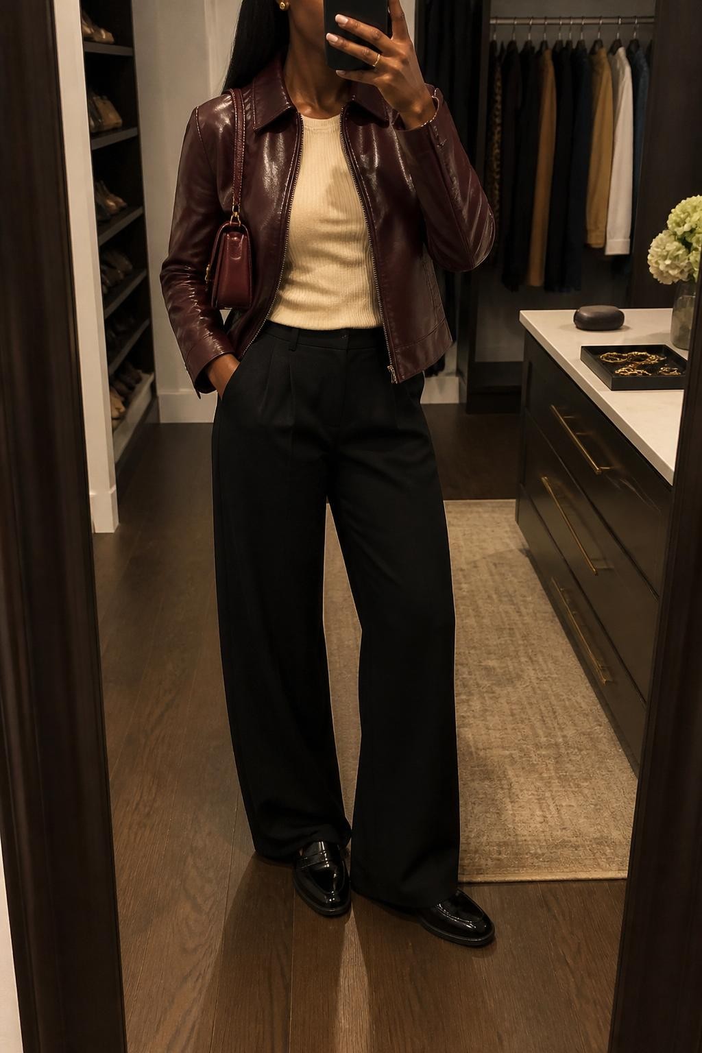 mirror selfie of a Black woman wearing the exact outfit described above, clearly showing a burgundy leather jacket layered over a cream ribbed knit top, black wide-leg trousers, black loafers, a burgundy mini handbag, and small gold stud earrings, holding a phone in front of a mirror, cropped at NECK so the face does not show, with a relaxed casual selfie pose, in a walk-in closet mirror, warm moody lighting, with an open closet with clothes visible and a jewelry tray on the vanity, latest iPhone photo quality.